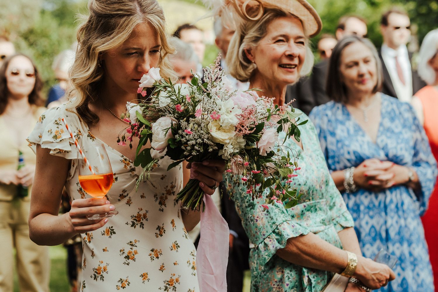 Wedding guests in floral dresses share drinks and laughter at an outdoor reception.
