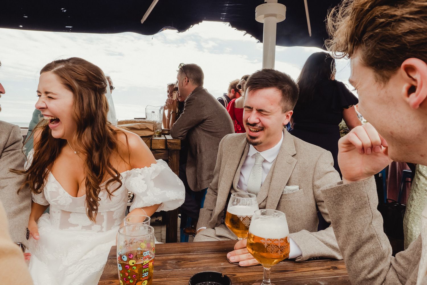 Wedding guests share a candid laughing moment while enjoying beers at an outdoor reception table.