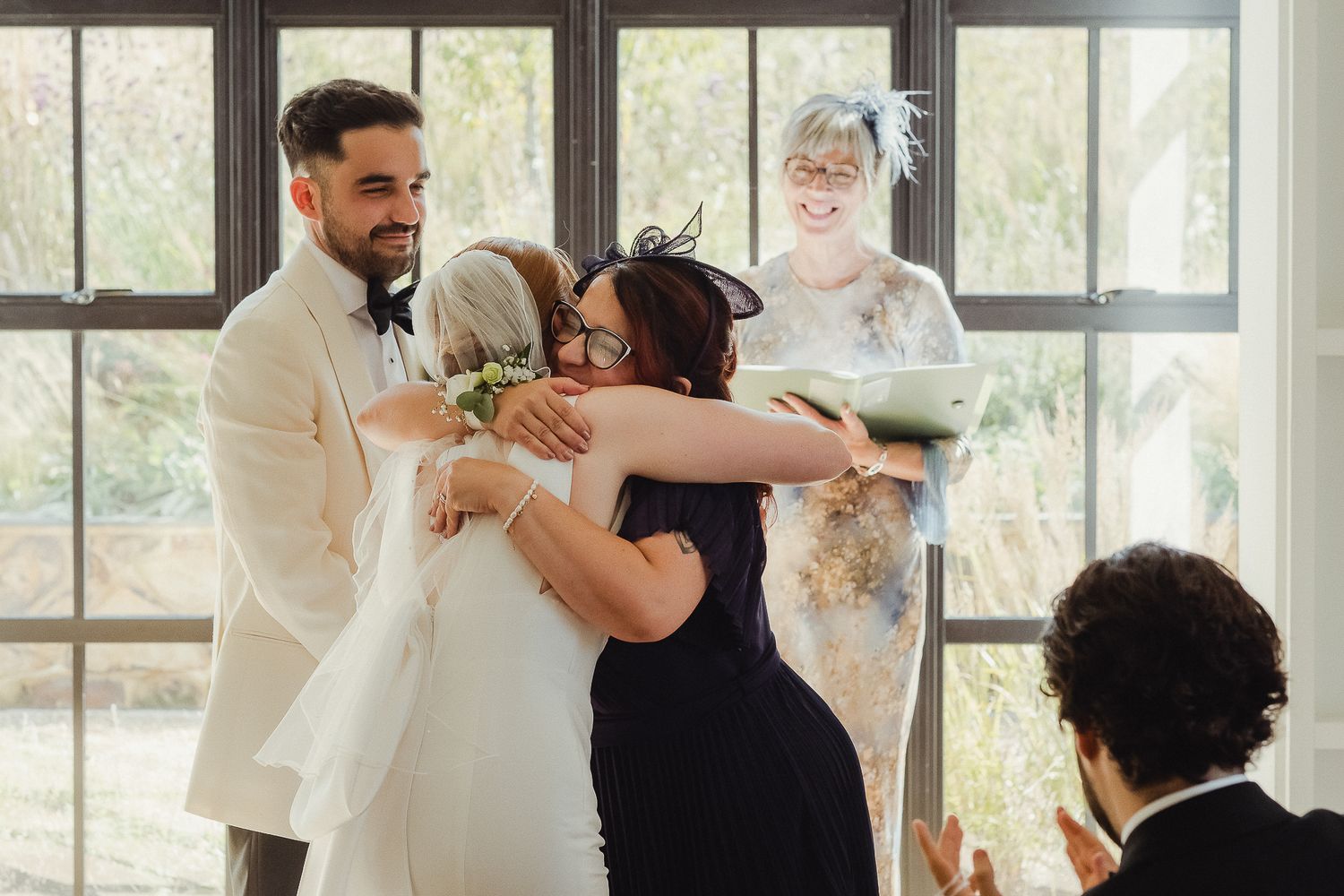 Wedding guests dance and celebrate together in a bright sunlit venue with large windows.