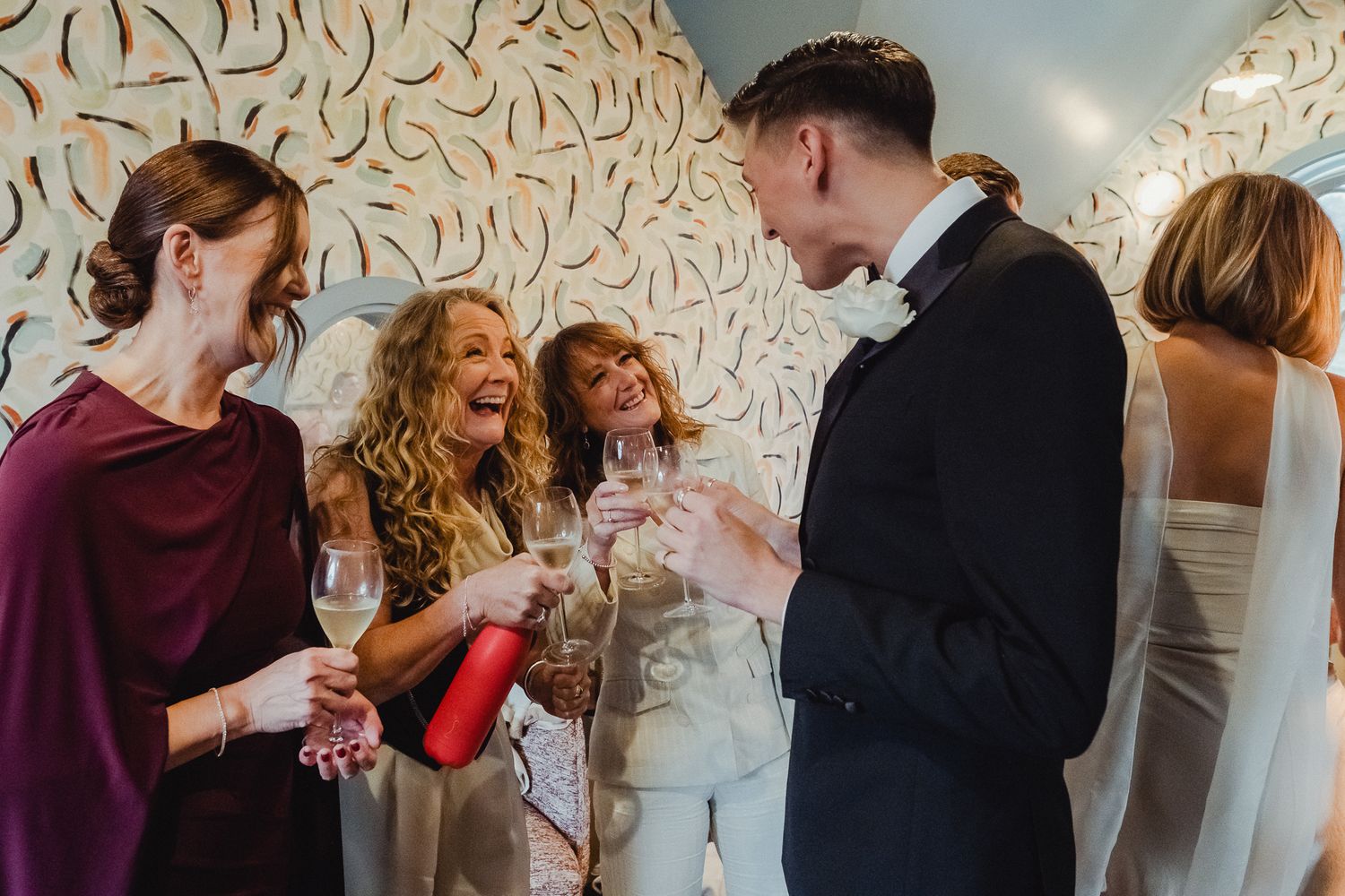 People dance and celebrate at a wedding reception in front of a decorative white wall with artistic swirl patterns.