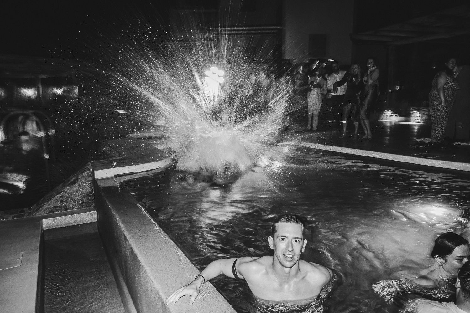 A bright explosion of sparks illuminates a swimming pool at night while swimmers watch from the water.