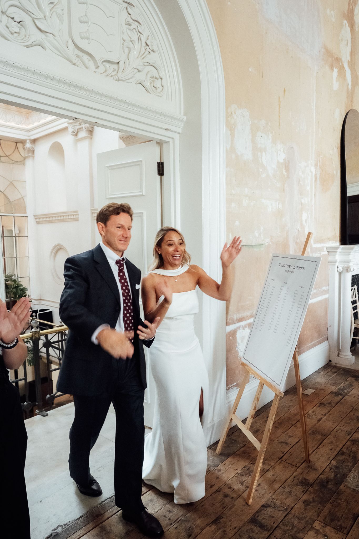 Bride in a white gown and groom in a navy suit enter a grand venue with peeling walls, waving as guests cheer beside a seating chart.