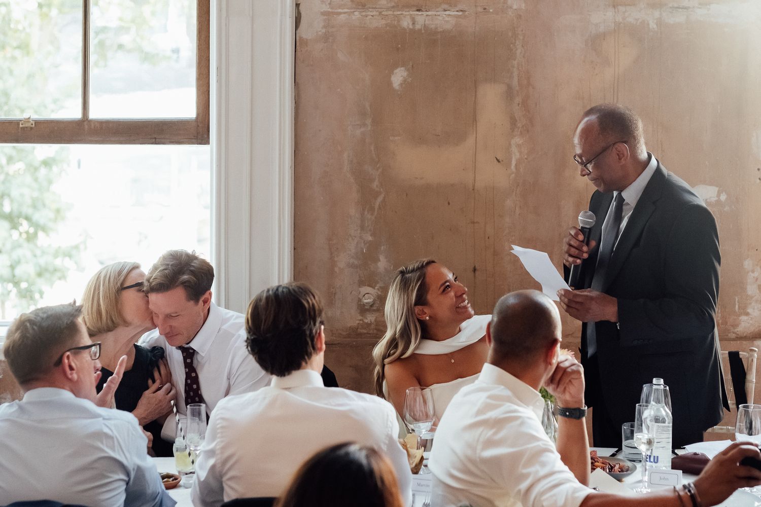 Man in a dark suit holds a microphone and reads from a paper while delivering a toast to seated wedding guests at a rustic reception.