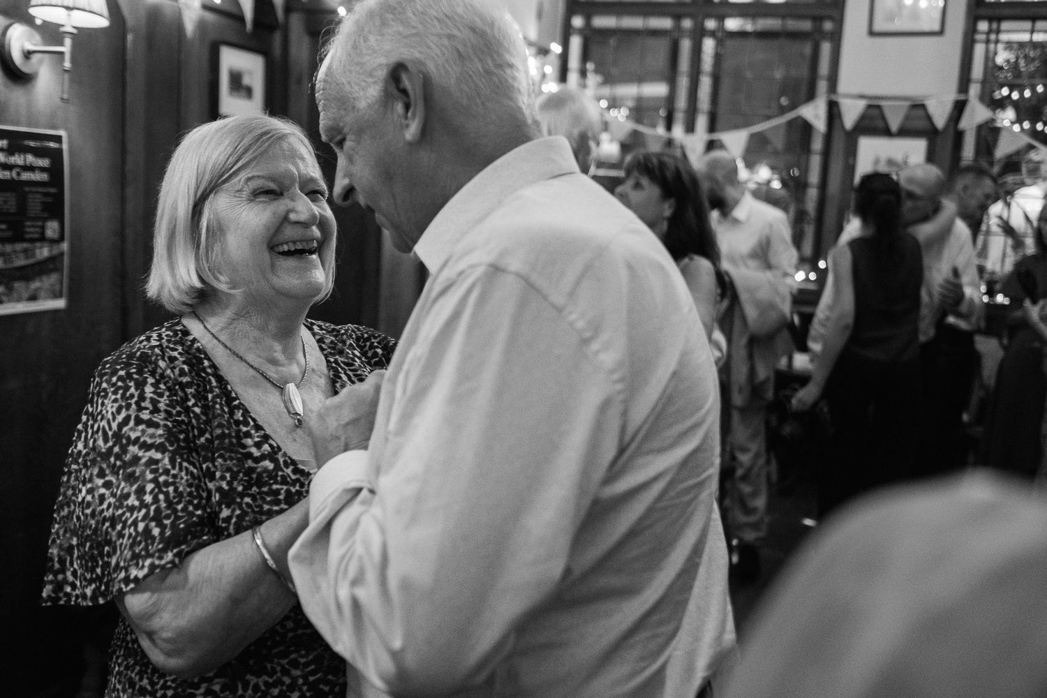 Smiling older woman in a patterned top dances closely with an older man in a white shirt at a lively indoor party with bunting.