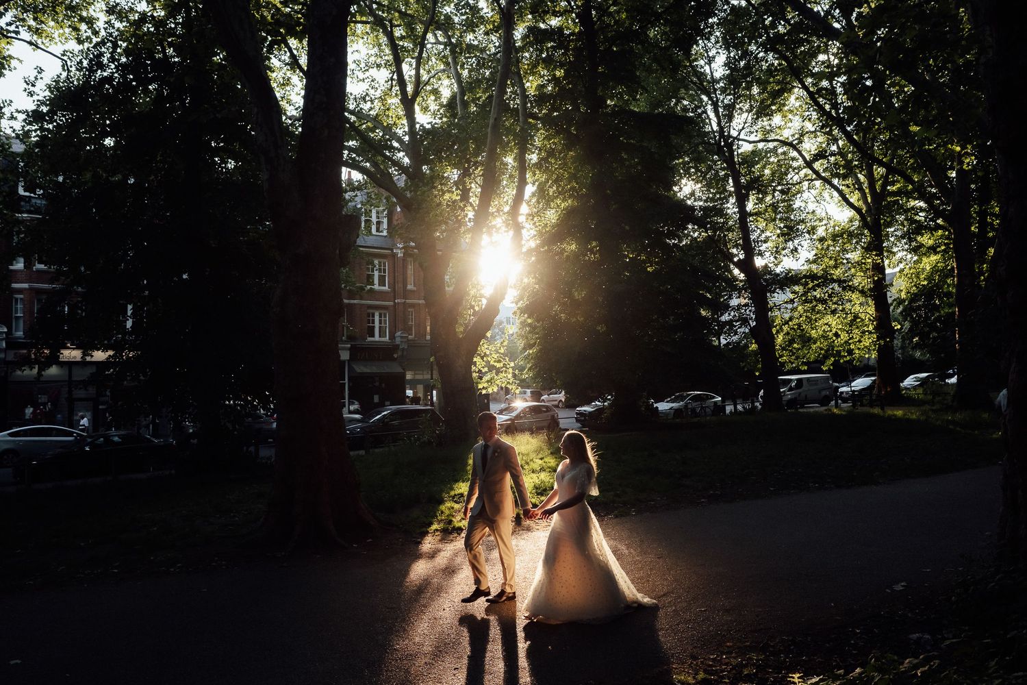 Bride in a white gown and groom walking hand in hand through a sunlit park path framed by tall trees at golden hour.