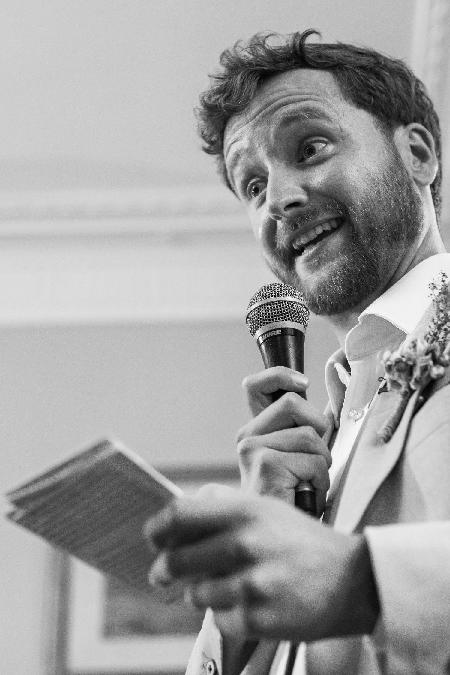 Bearded man with curly hair giving a wedding speech, holding a microphone and note cards, wearing a floral boutonniere.