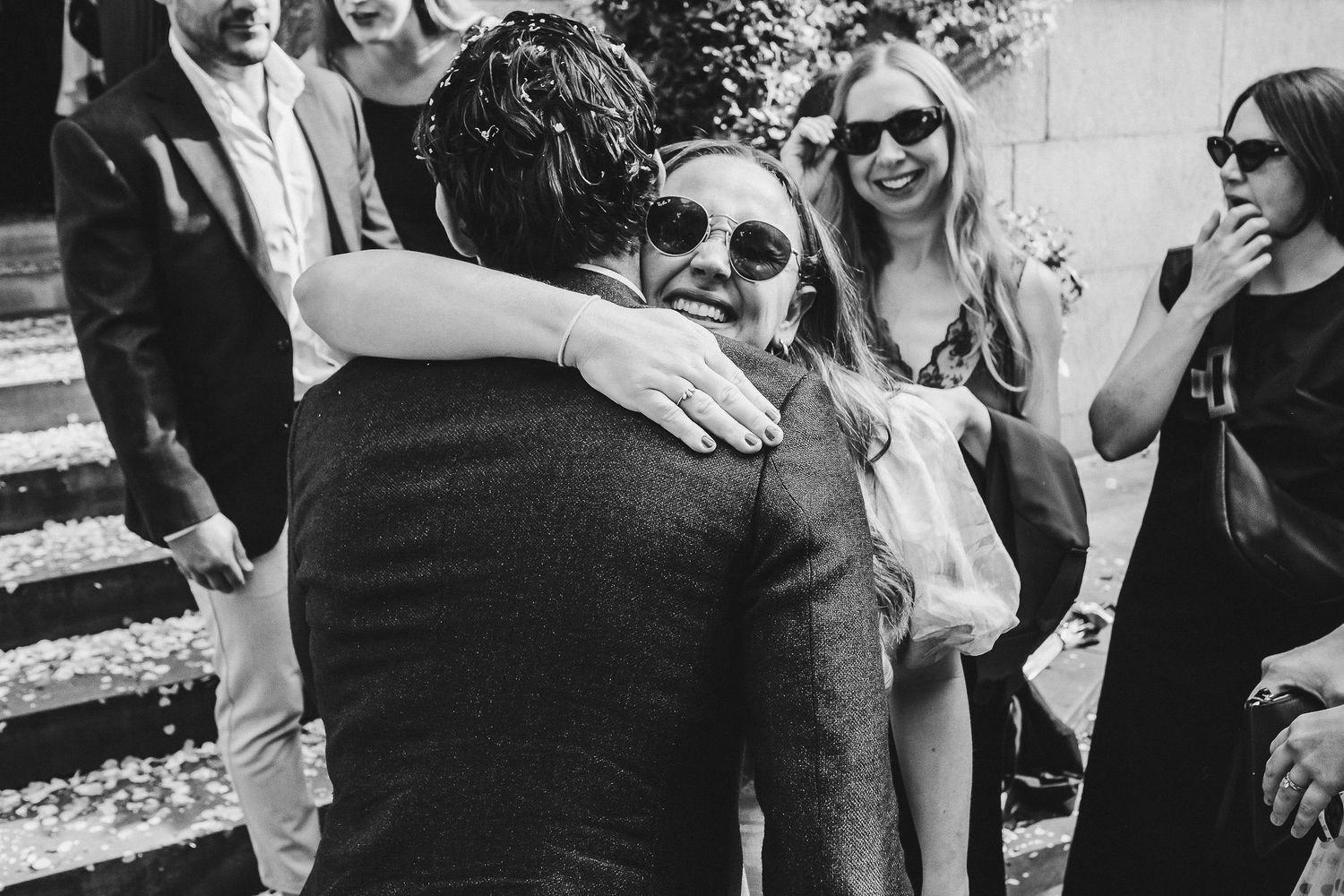 A candid black and white photograph showing people embracing and laughing together outdoors in sunglasses.