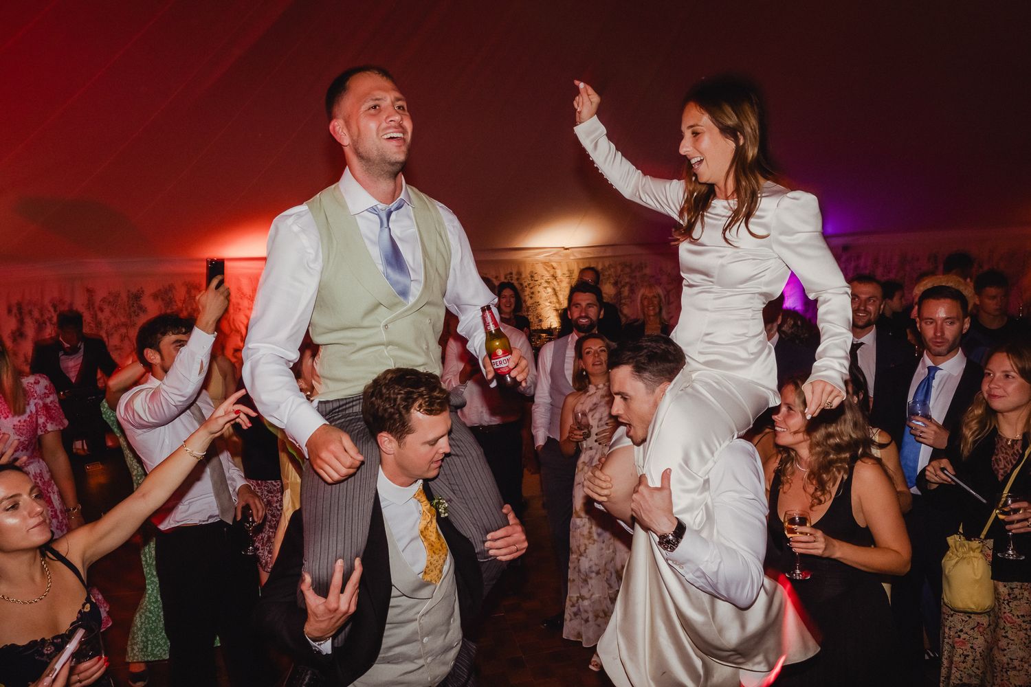 Wedding guests dance and celebrate on a dimly lit dance floor with red and purple lighting effects.