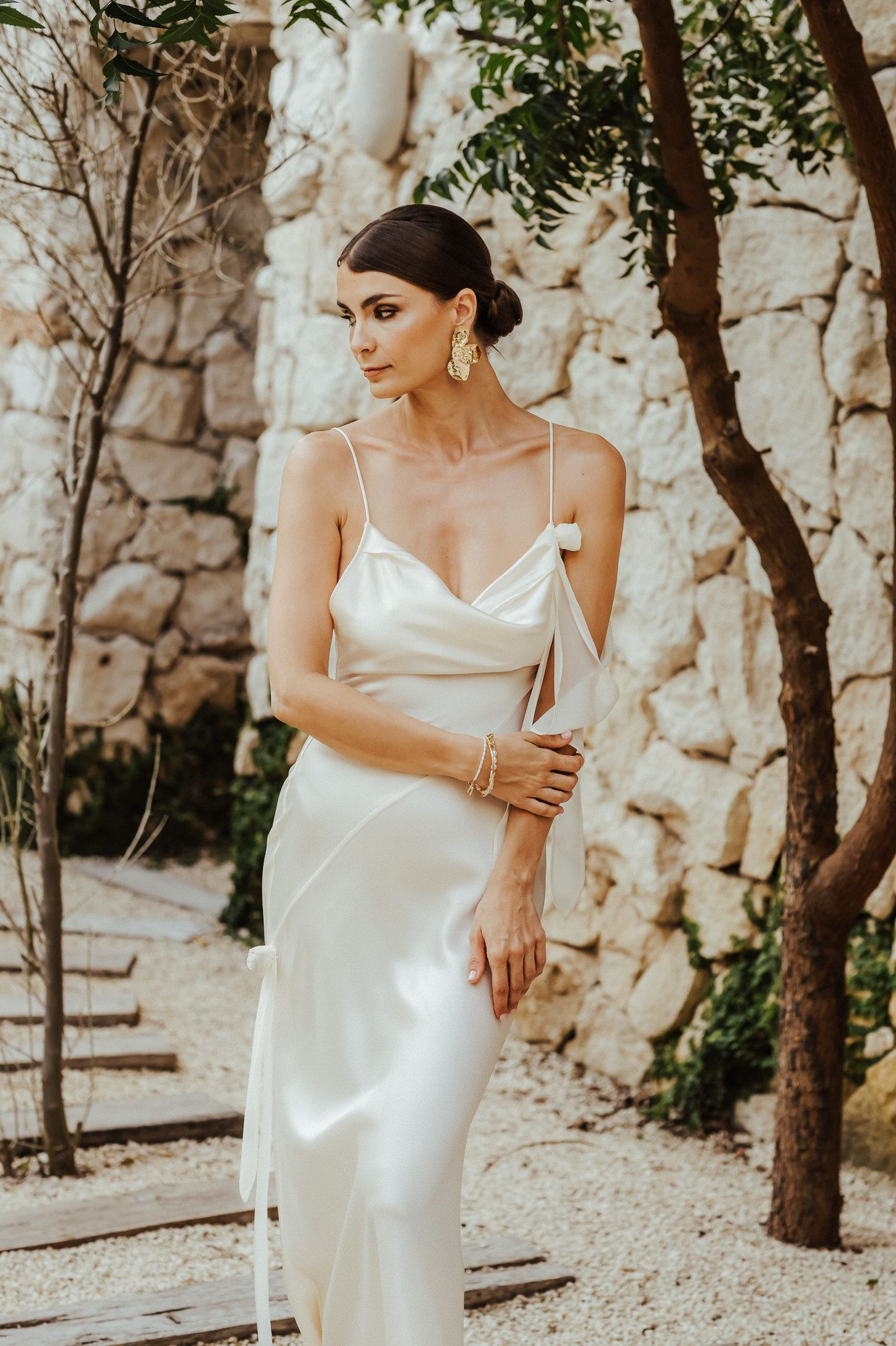 A bride in a silky white slip dress poses elegantly against a rustic stone wall in an outdoor garden setting.