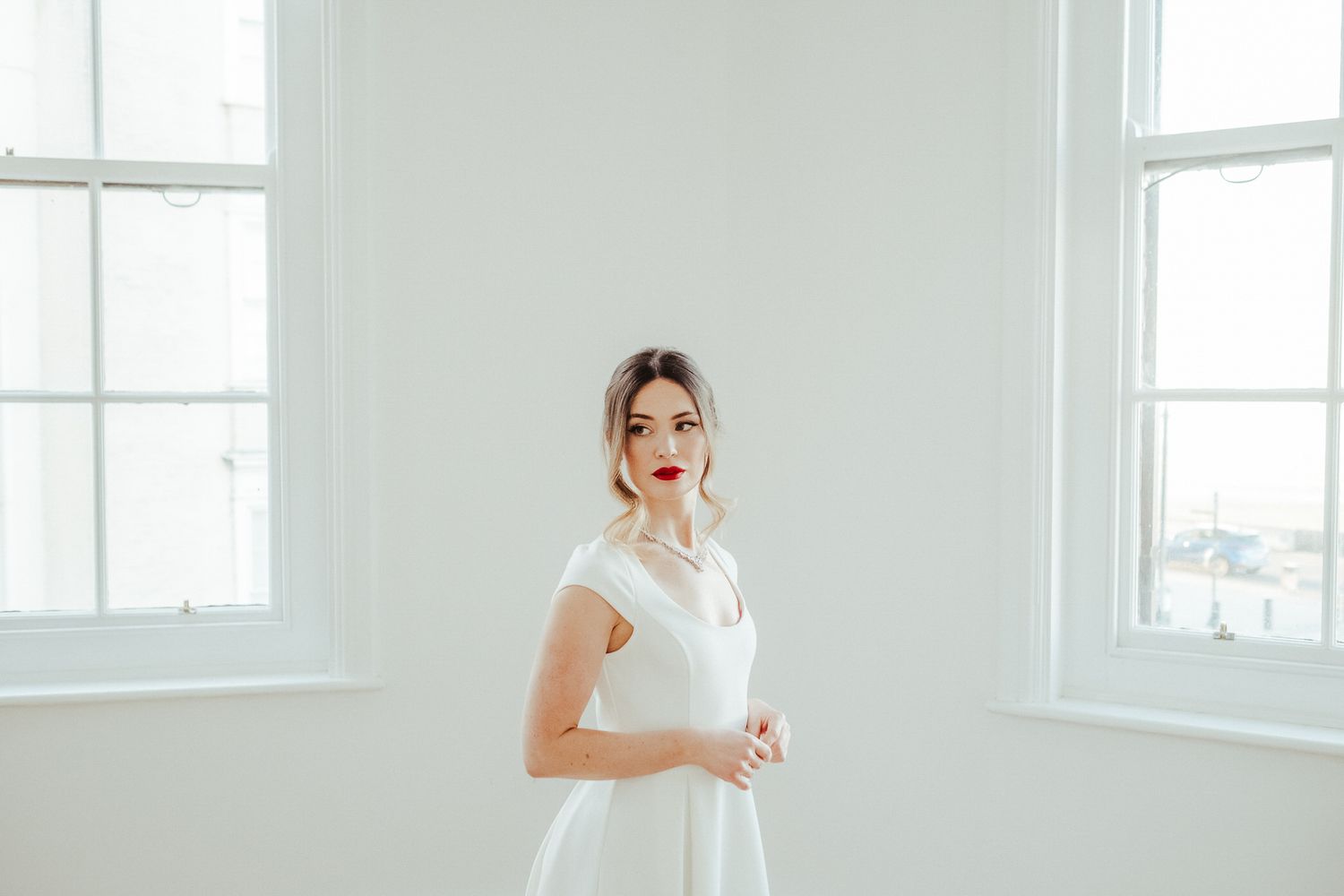 A person in a white wedding dress poses gracefully by bright windows in an elegant white room.