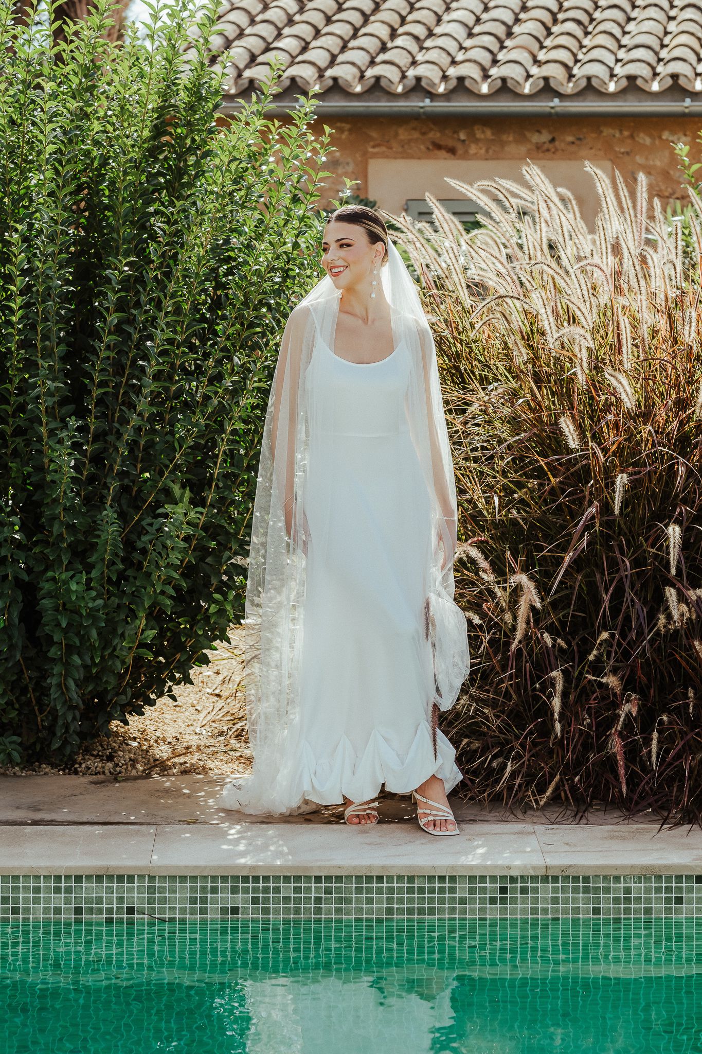 A bride in a flowing white dress sits elegantly beside a turquoise swimming pool with lush greenery in the background.