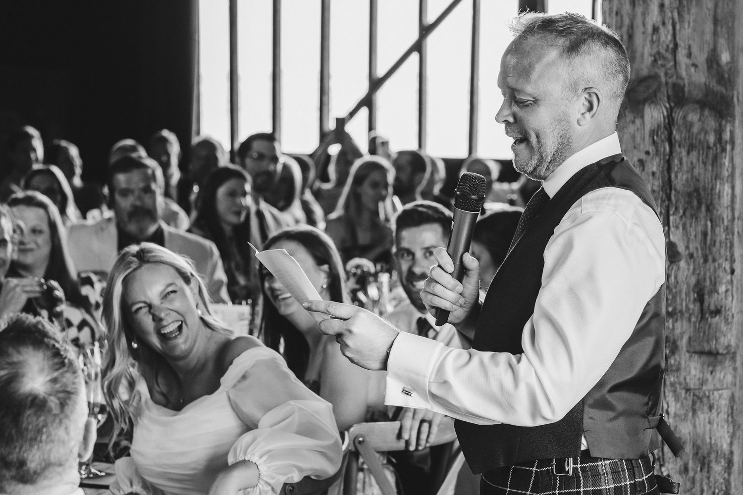 Wedding guests laugh and smile during a heartfelt reception speech in a rustic barn venue.