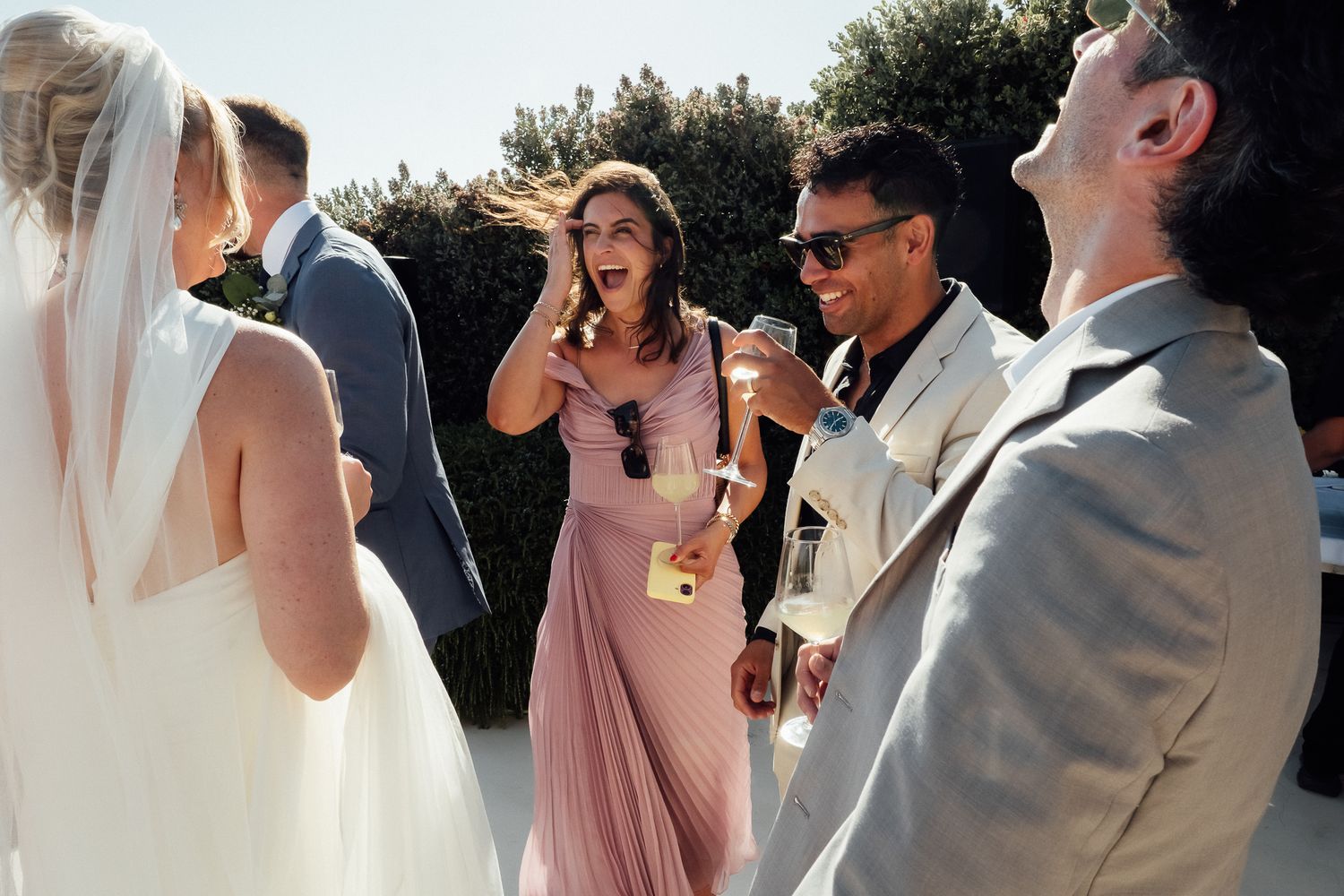 Wedding guests in elegant attire socializing outdoors, bridesmaid in pink dress conversing with guests on sunny day.