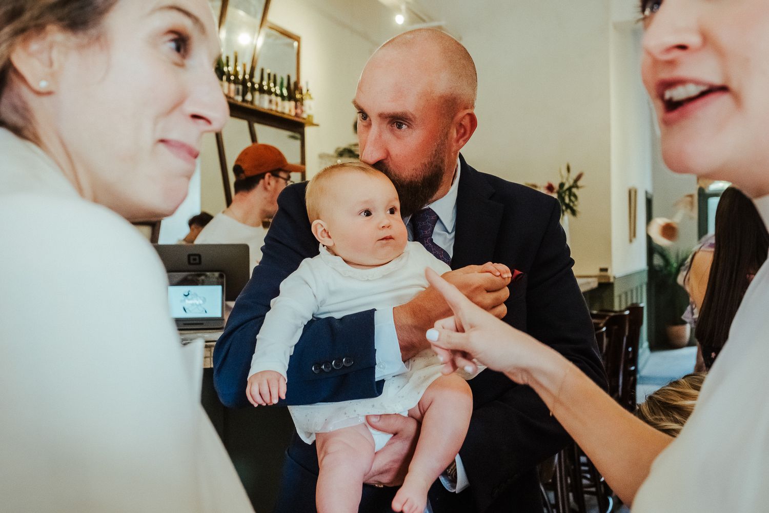 A baby in a white outfit surrounded by adults in an indoor setting with soft natural lighting.