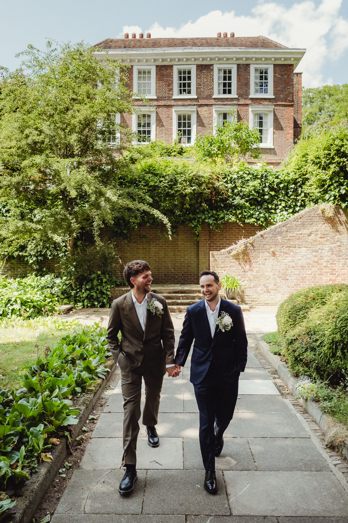 Two grooms walking hand in hand along a garden path in front of a Georgian brick manor house.
