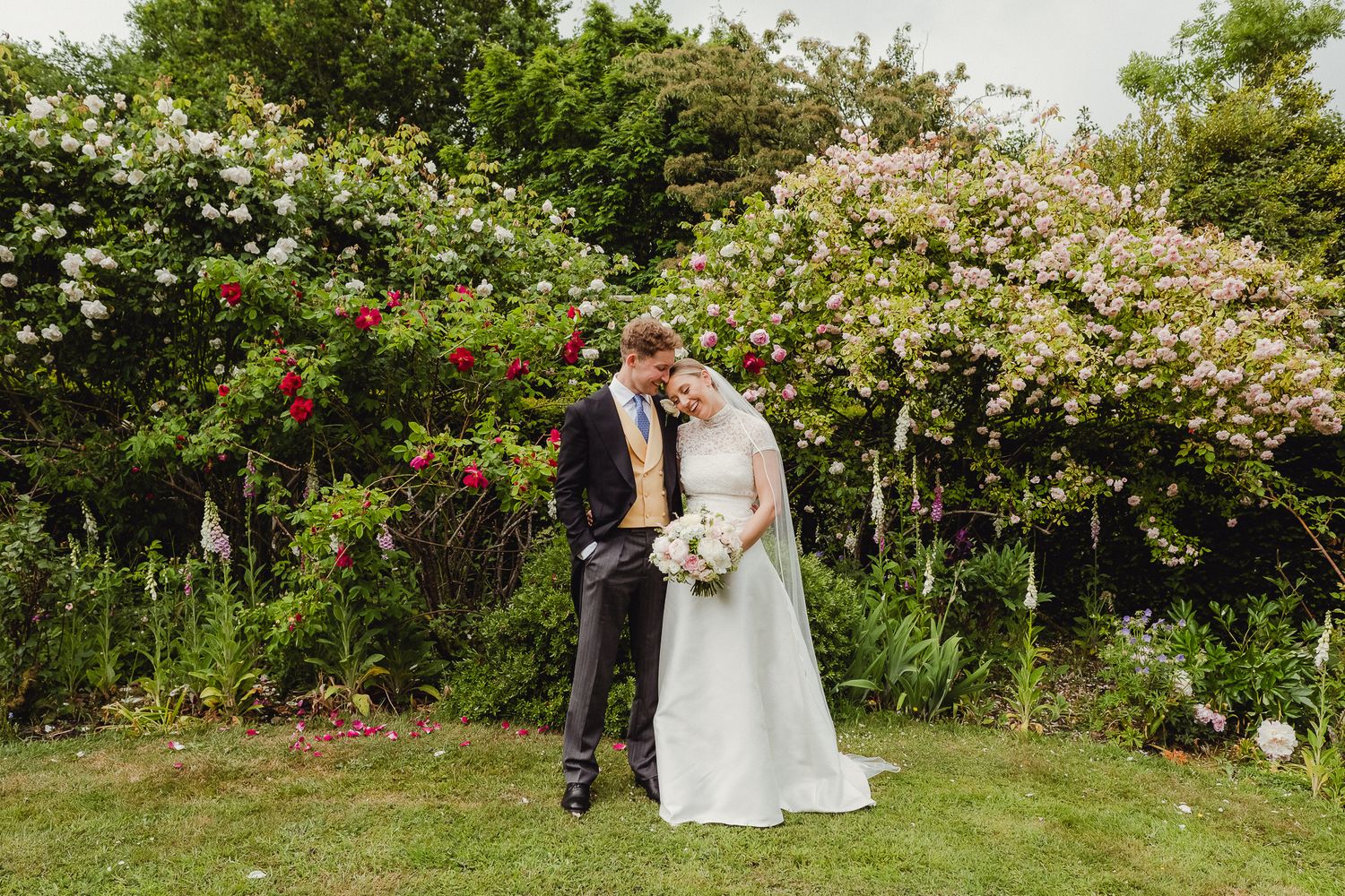 A couple shares an intimate moment surrounded by lush greenery and blooming flowers in a garden setting.