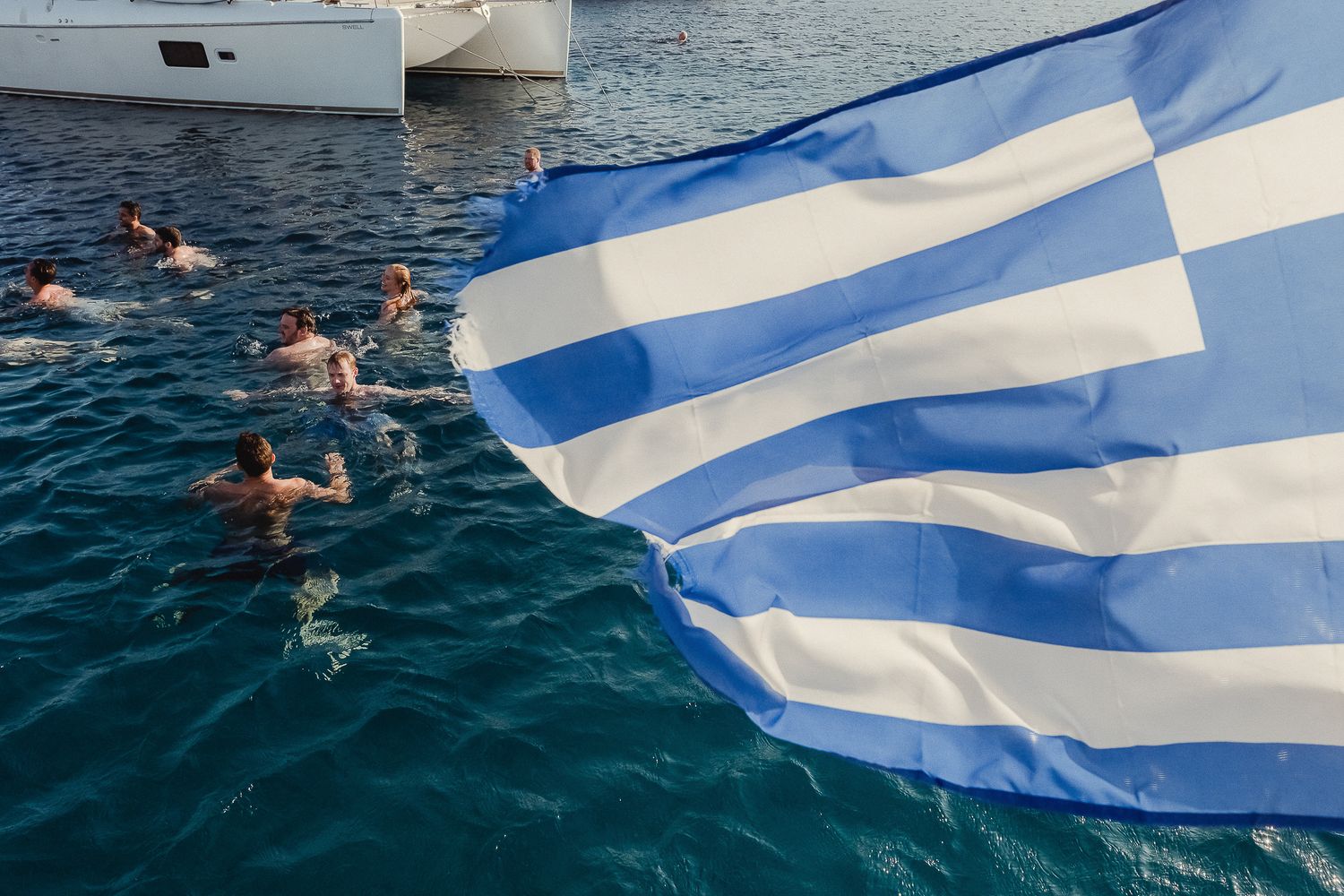 A series of photos showing someone swimming in blue ocean waters while waving a Greek flag against a bright sunny sky.