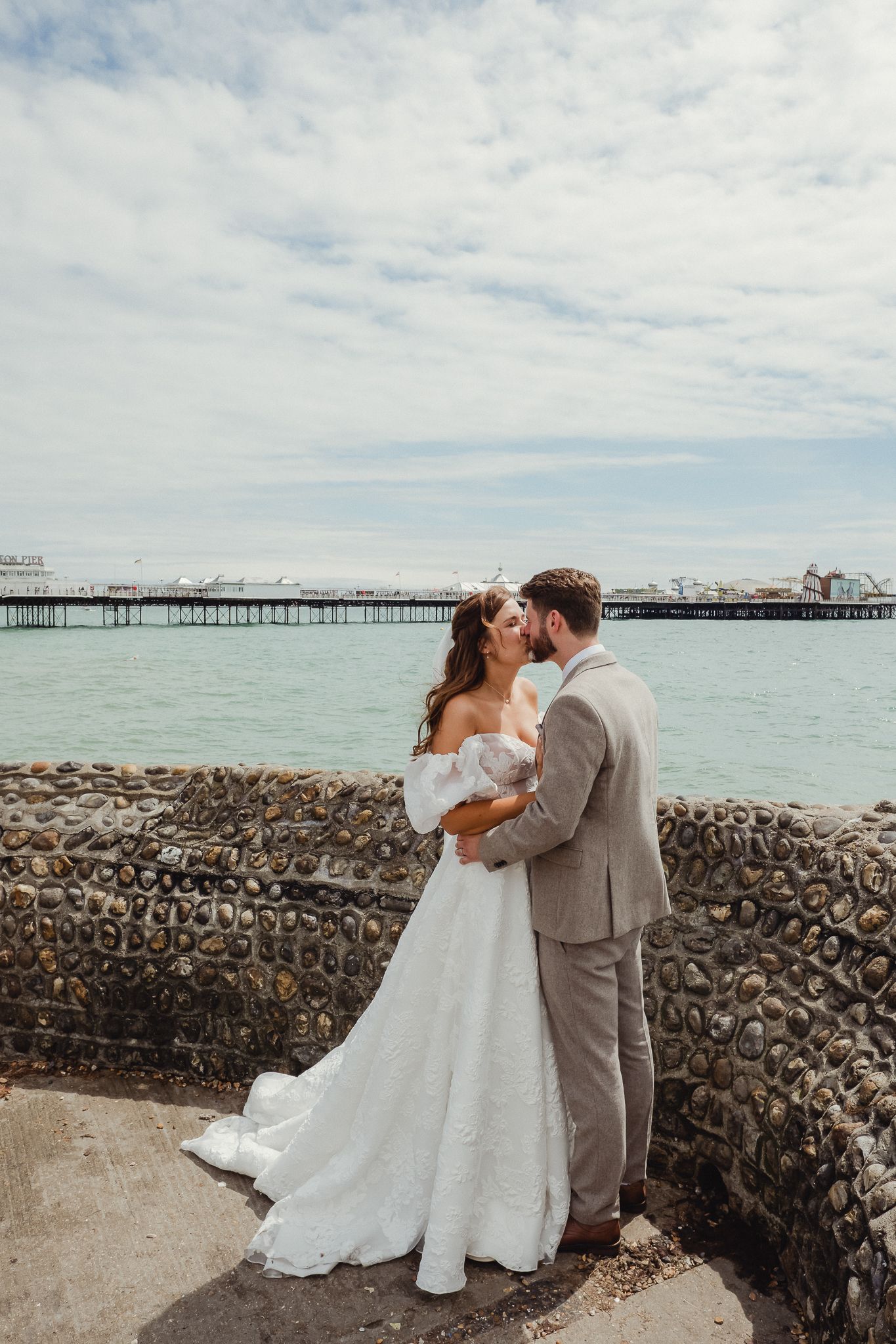 A romantic seaside wedding moment captured against a stone wall with an ocean pier visible in the background.