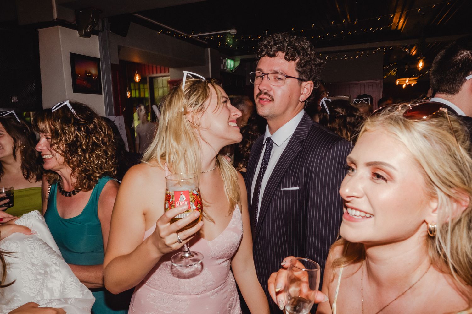 Wedding reception guests dance and celebrate on a crowded dance floor with party lights in the background.