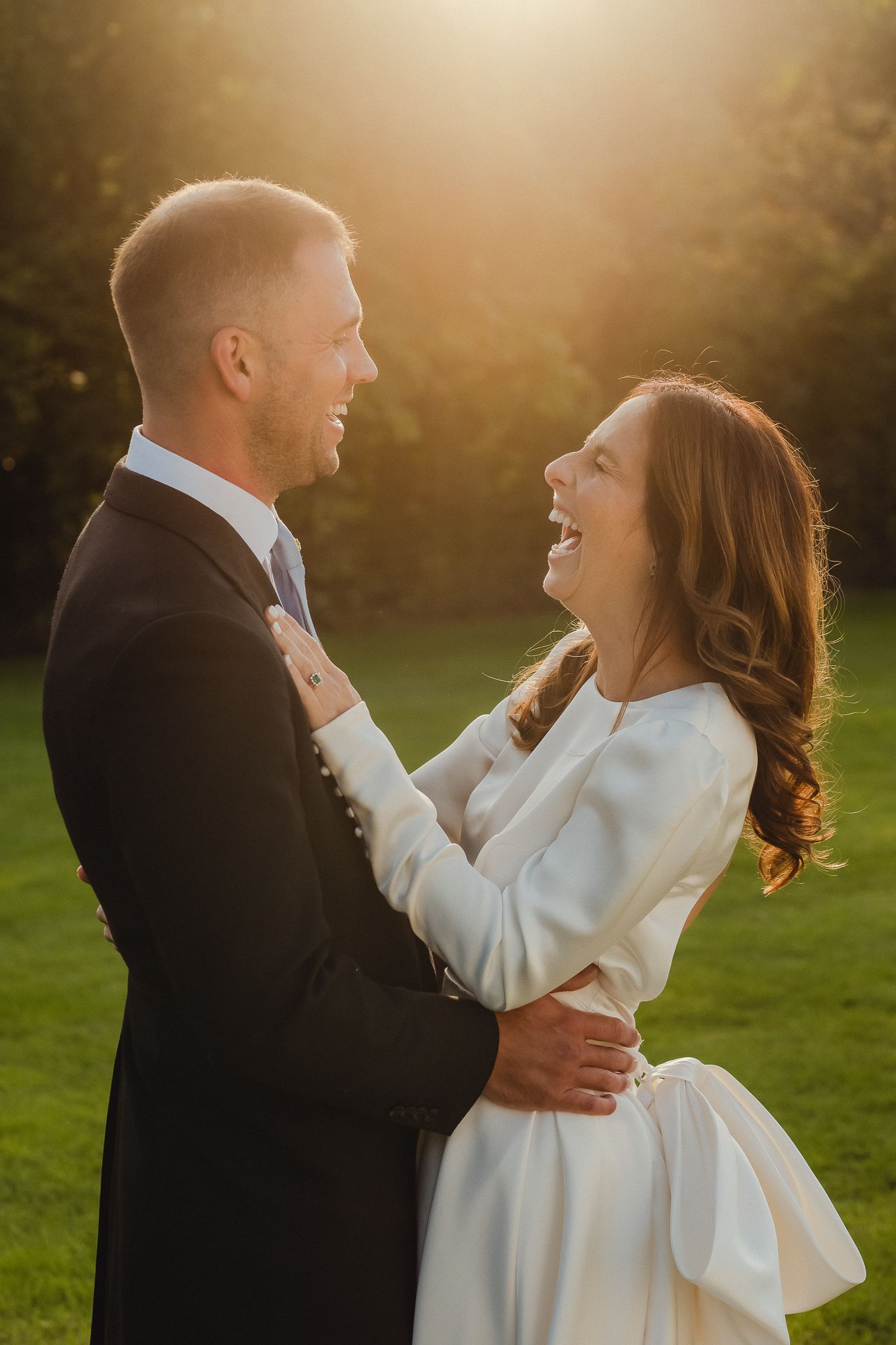 A couple shares a joyful moment in a sunlit garden, with the bride wearing an elegant white long-sleeved dress.