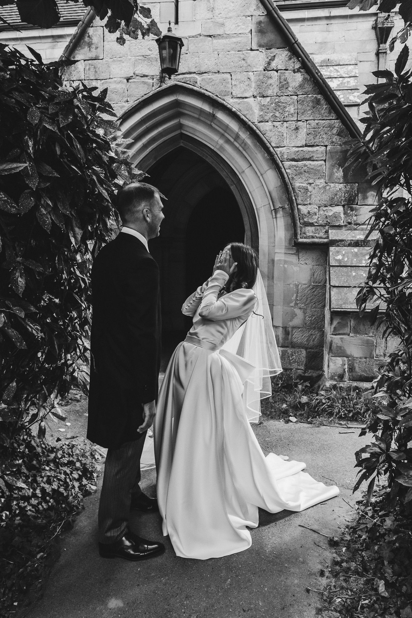 A black and white wedding photo showing a couple embracing in front of a gothic stone church archway surrounded by foliage.