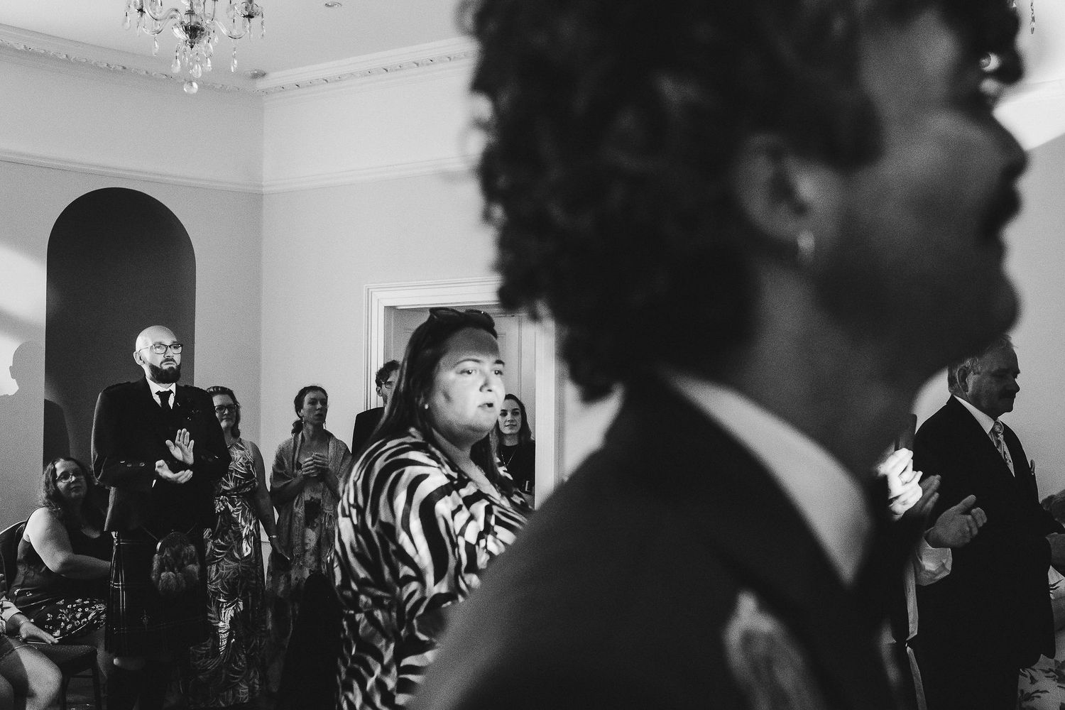 Black and white photo of wedding guests standing in an elegant room, with a woman in a zebra-print top watching the ceremony beside a man in