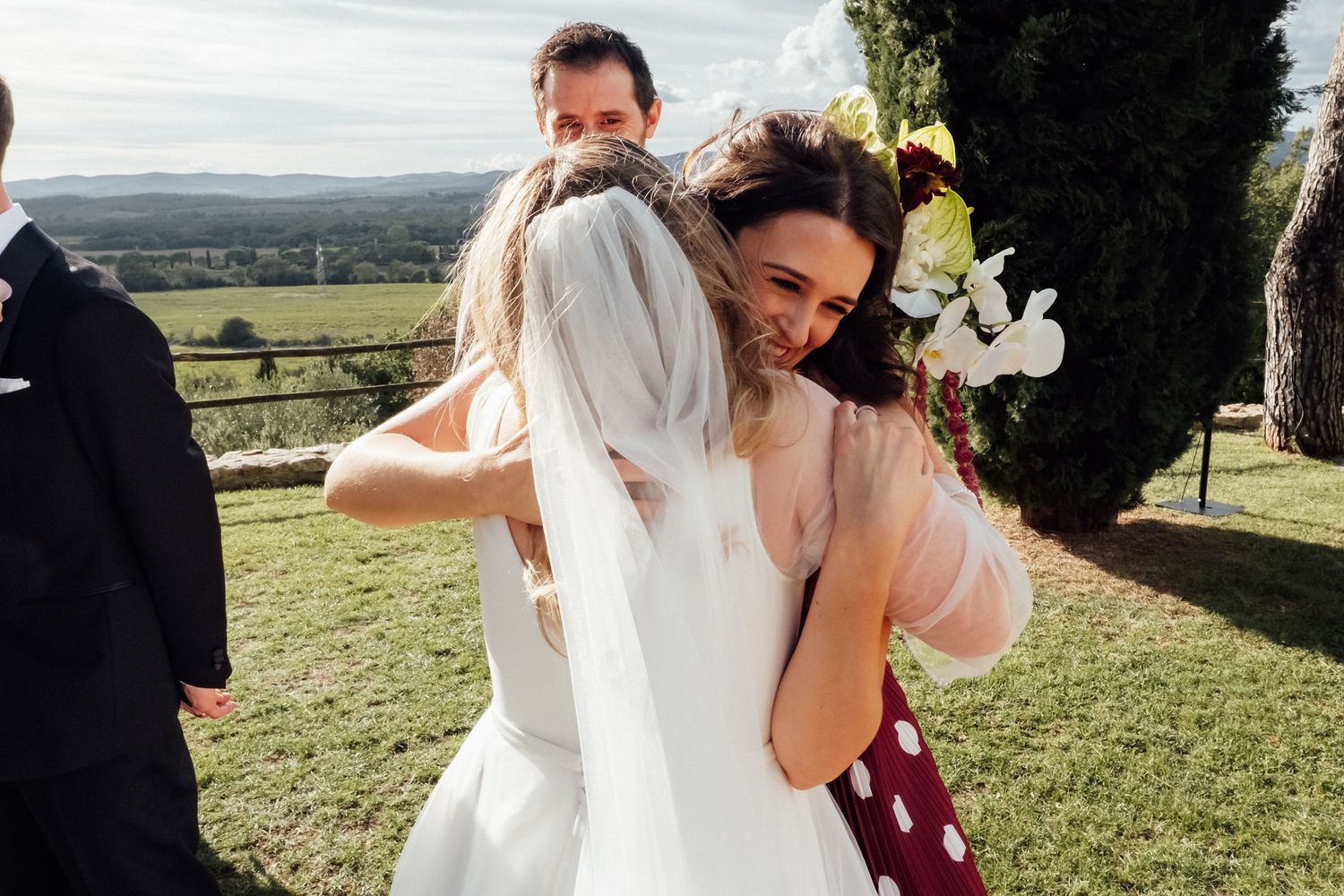 Bride in a white veil and gown embraces a smiling guest in a polka dot dress at an outdoor wedding with green hills behind.