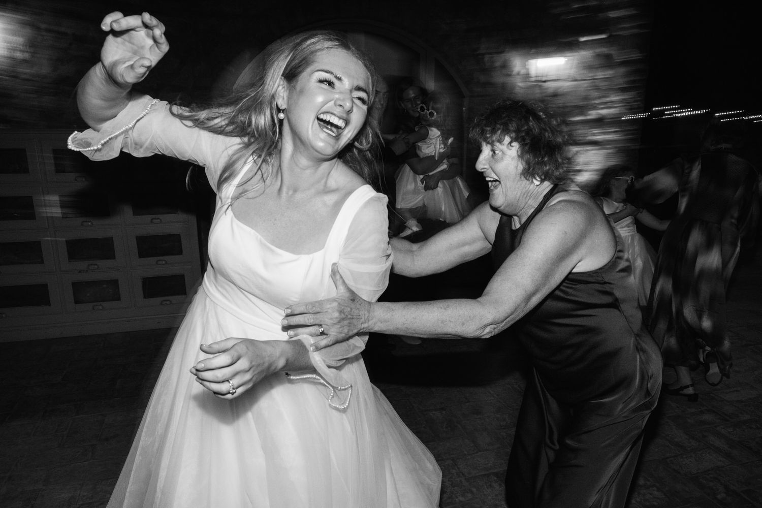 Black and white photo of a laughing bride in a white gown dancing joyfully with a smiling older woman at a lively wedding reception.