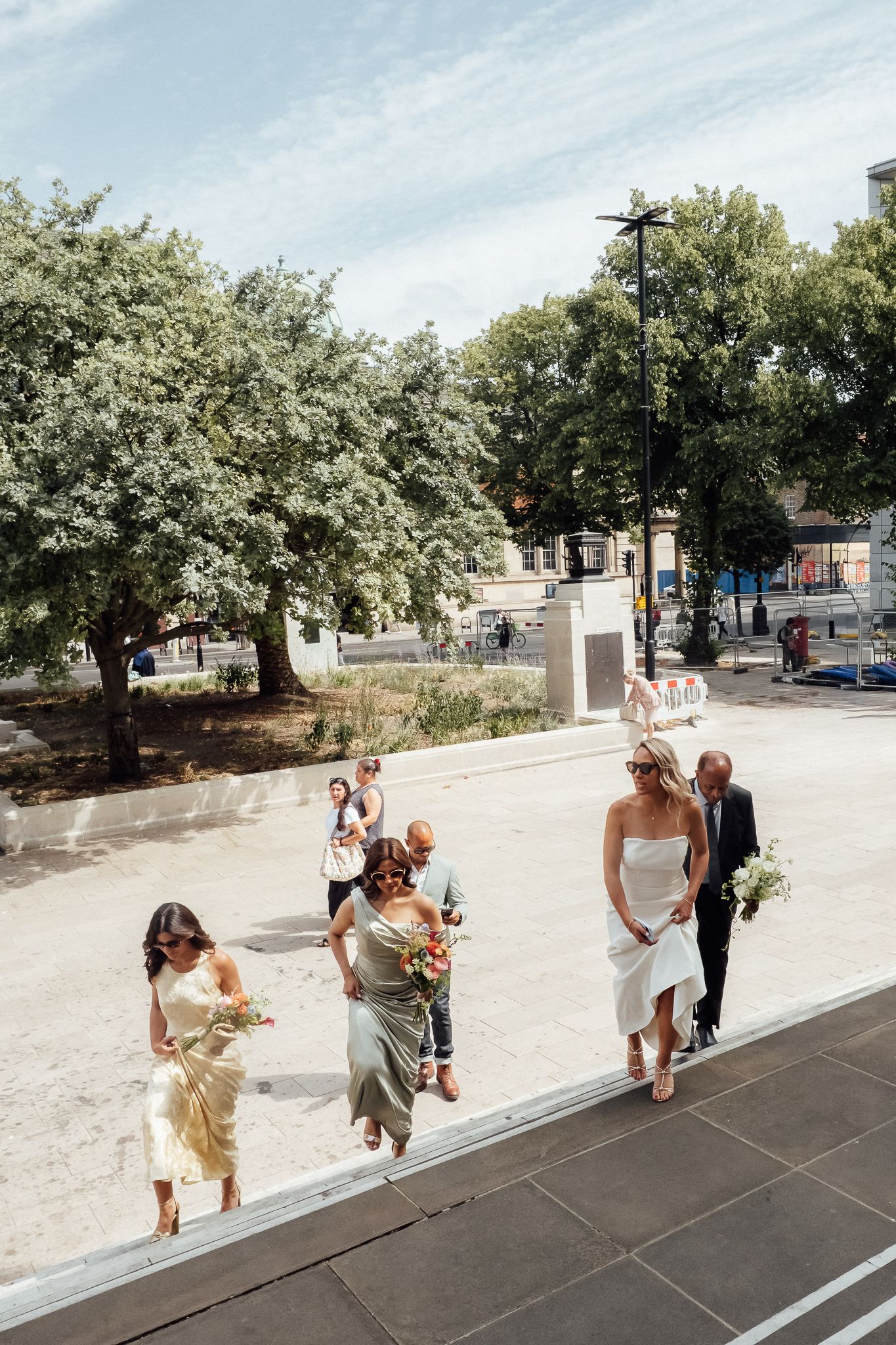 Bride in a white dress carrying flowers walks with a man in a suit as bridesmaids in satin gowns approach a building on a sunny day.