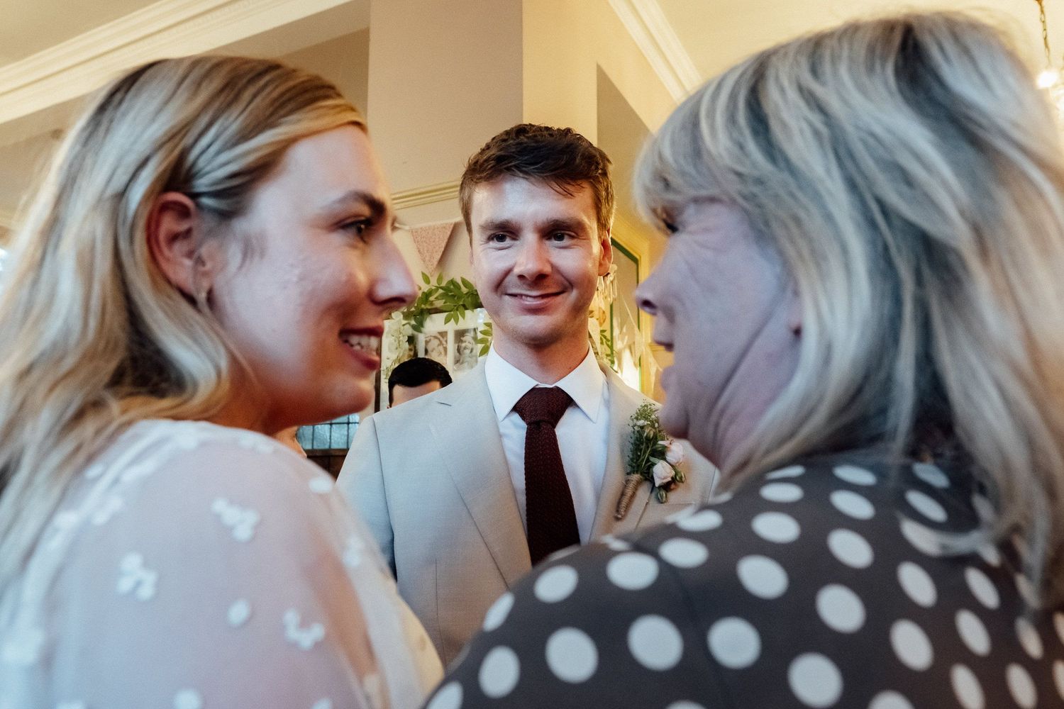 Smiling young man in a gray suit and burgundy tie with a boutonniere chats with two women at a festive indoor celebration.