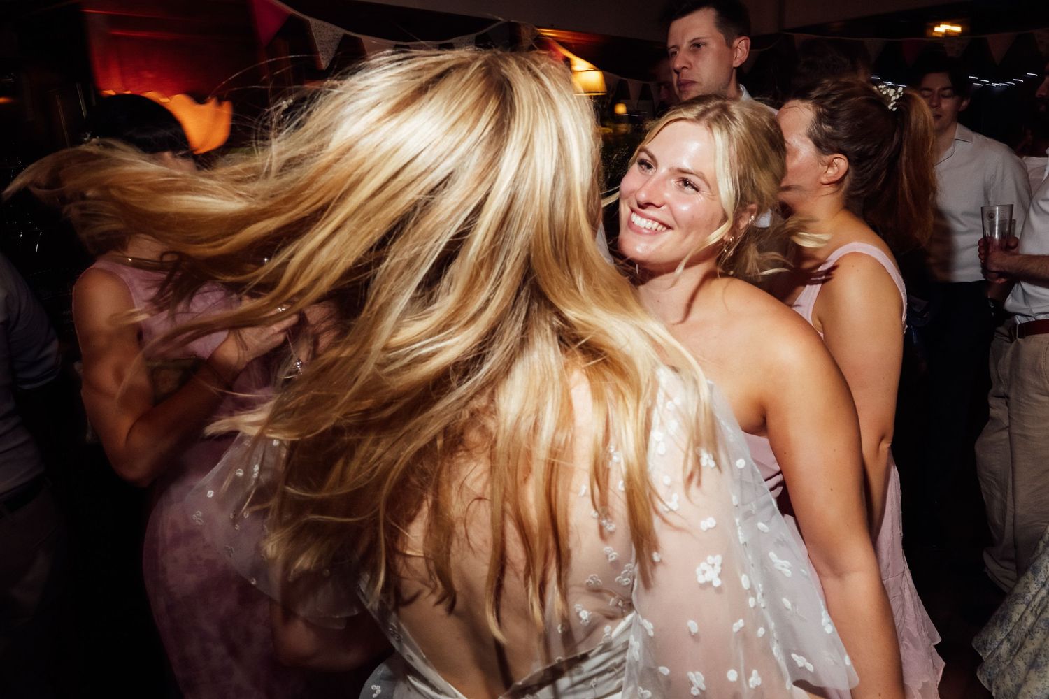 Smiling woman in a white dress dances at a lively party while a blonde woman's hair swings in motion in the foreground.
