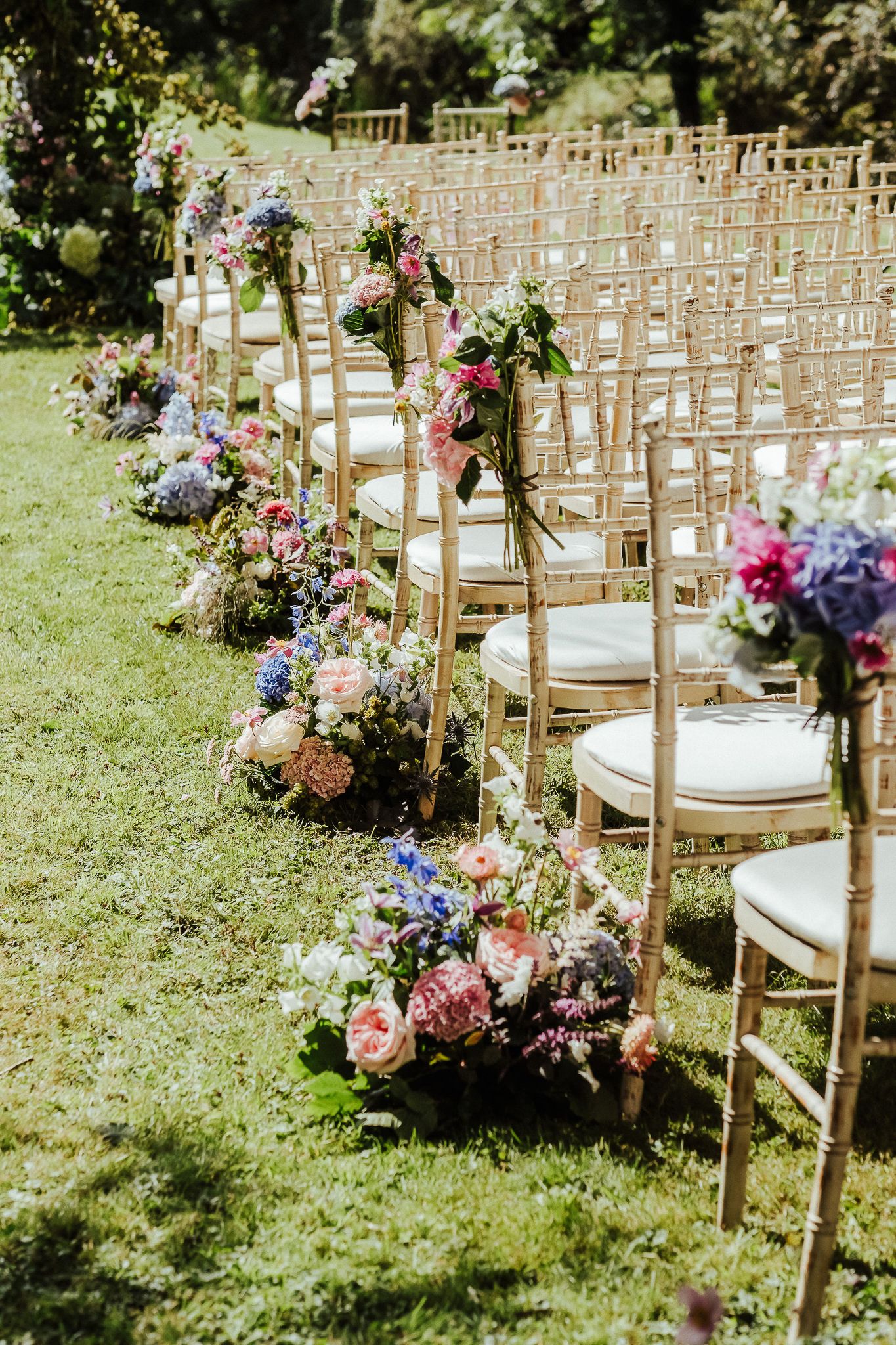 Gold chairs lined up for an outdoor wedding ceremony with colorful floral aisle decorations.