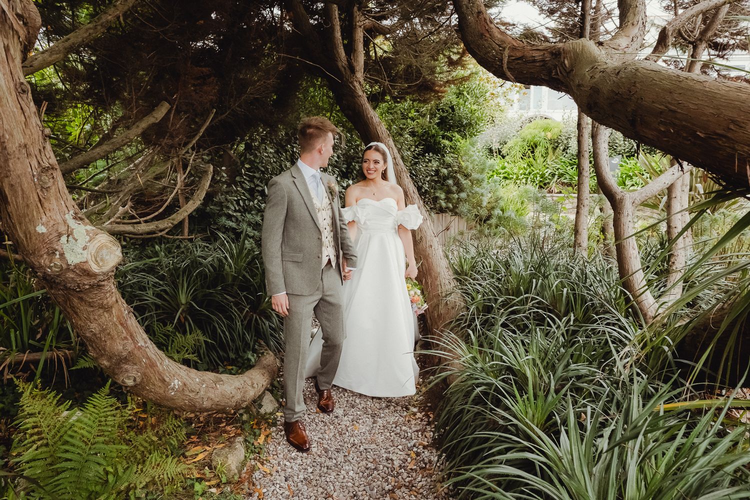 A couple walks through a lush garden pathway surrounded by tropical plants and twisted tree branches.