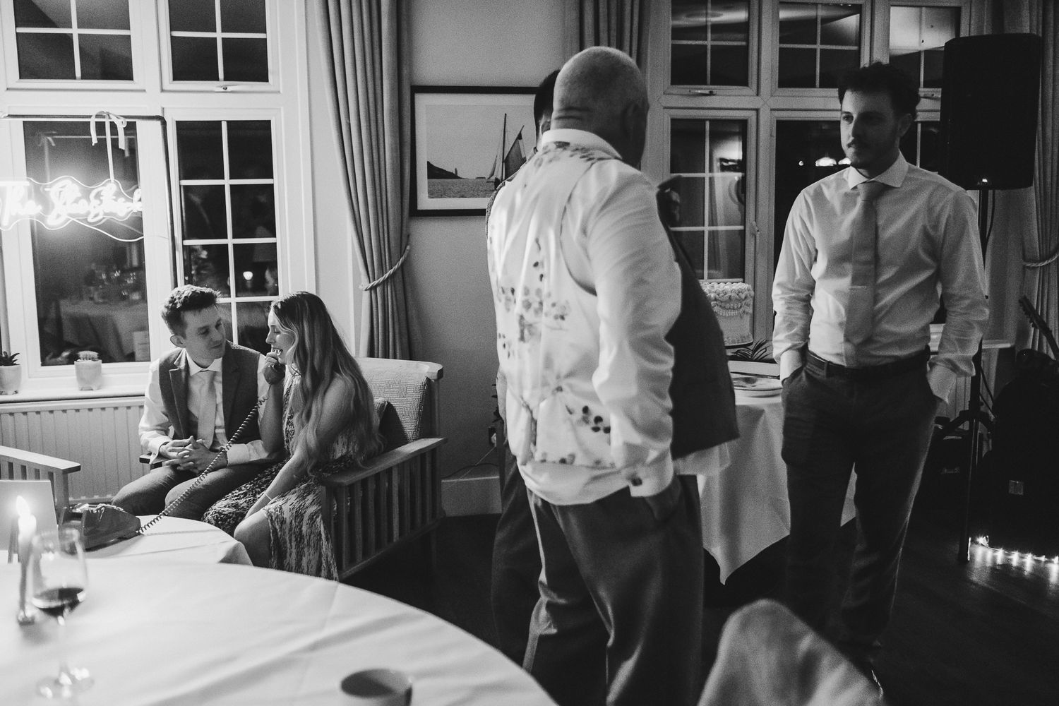 Black and white photo of guests mingling at an indoor event with windows and curtains in the background.