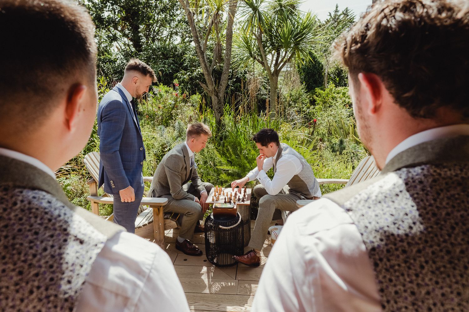 People in formal attire gather around a chess game outdoors in a garden setting.