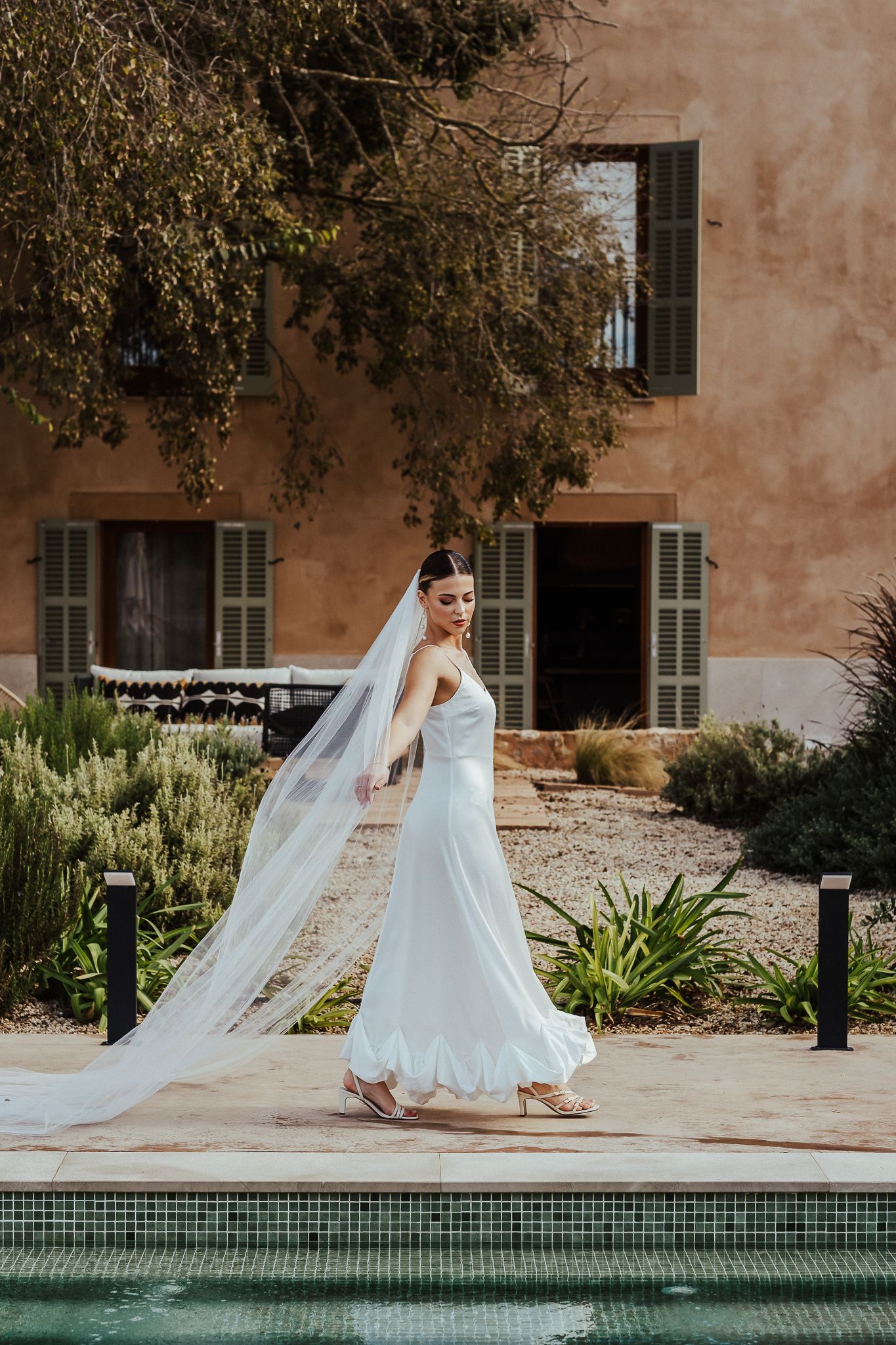 Elegant wedding dress and flowing veil catch the sunlight by a Mediterranean-style pool and terracotta building.