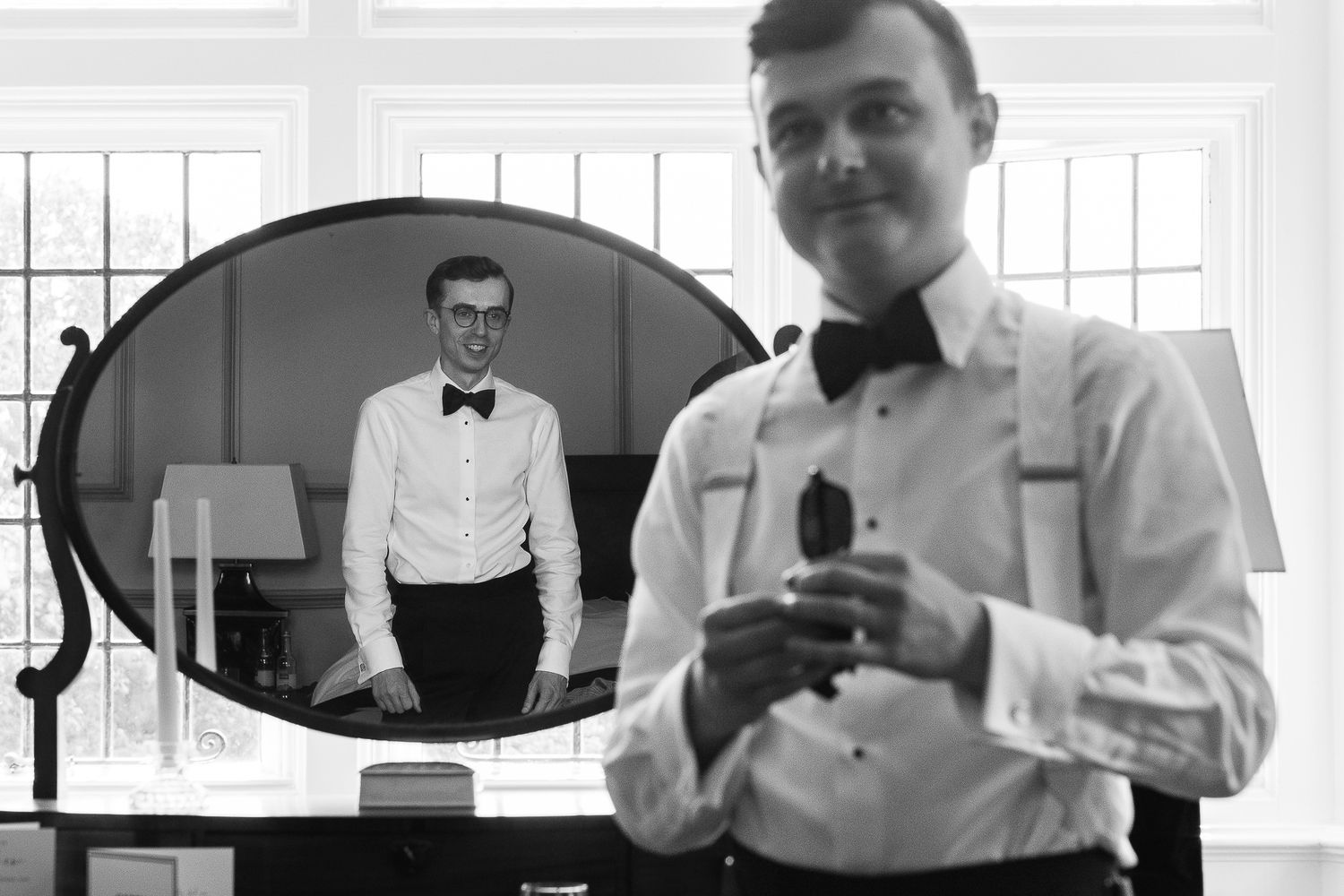 Black and white photo of a man in bow tie and suspenders holding glasses, reflected in an oval mirror.