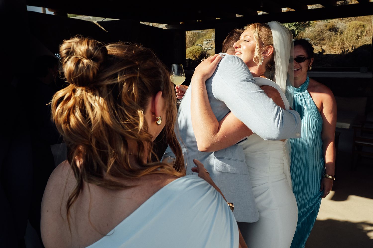 Bridesmaids in blue dresses share warm hugs at an outdoor wedding celebration in bright sunlight.