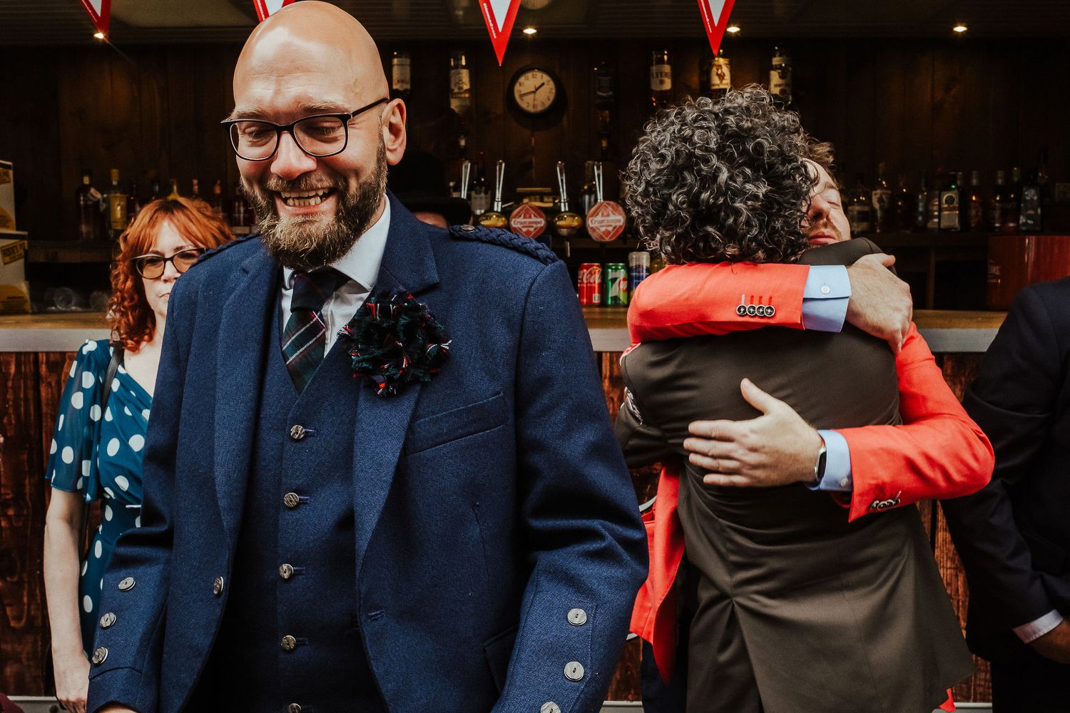 Smiling bald bearded man in glasses wearing a navy tweed suit with a floral boutonniere as two guests embrace in a cozy pub.