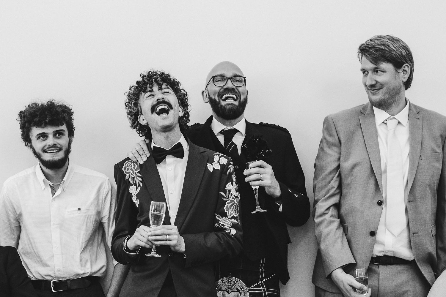 Four laughing men in formal wedding attire stand against a plain wall, two holding champagne glasses, in a joyful black and white photo.