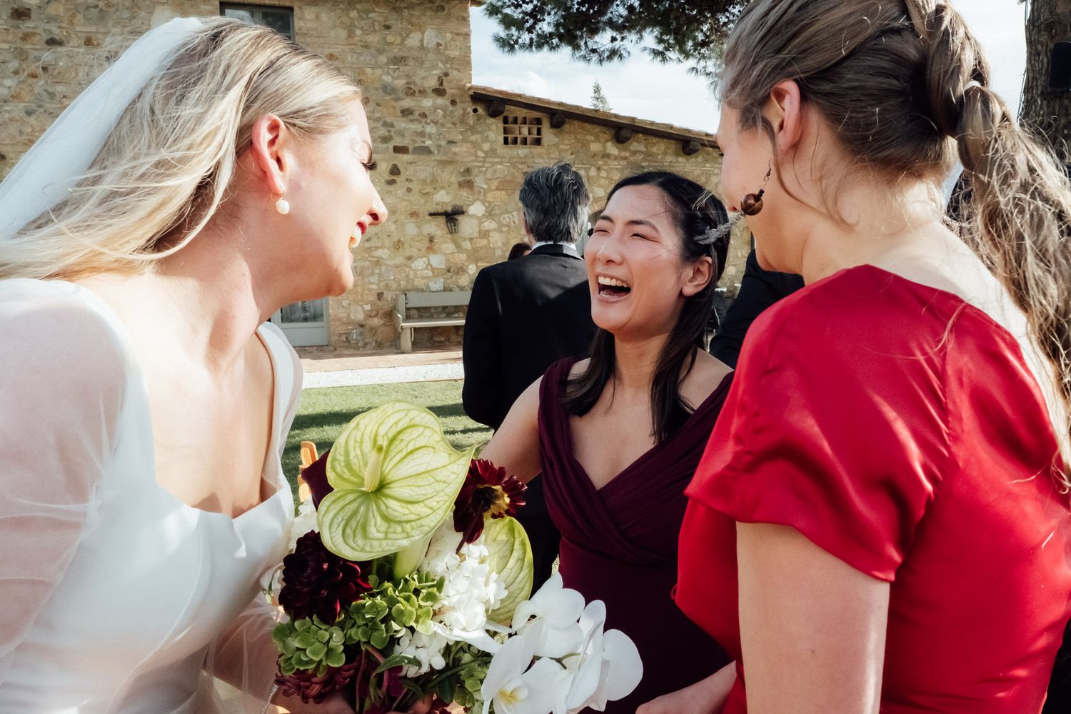 Three women laugh and chat at an outdoor wedding, with a bride in a veil and a bouquet of white and green flowers in the foreground.