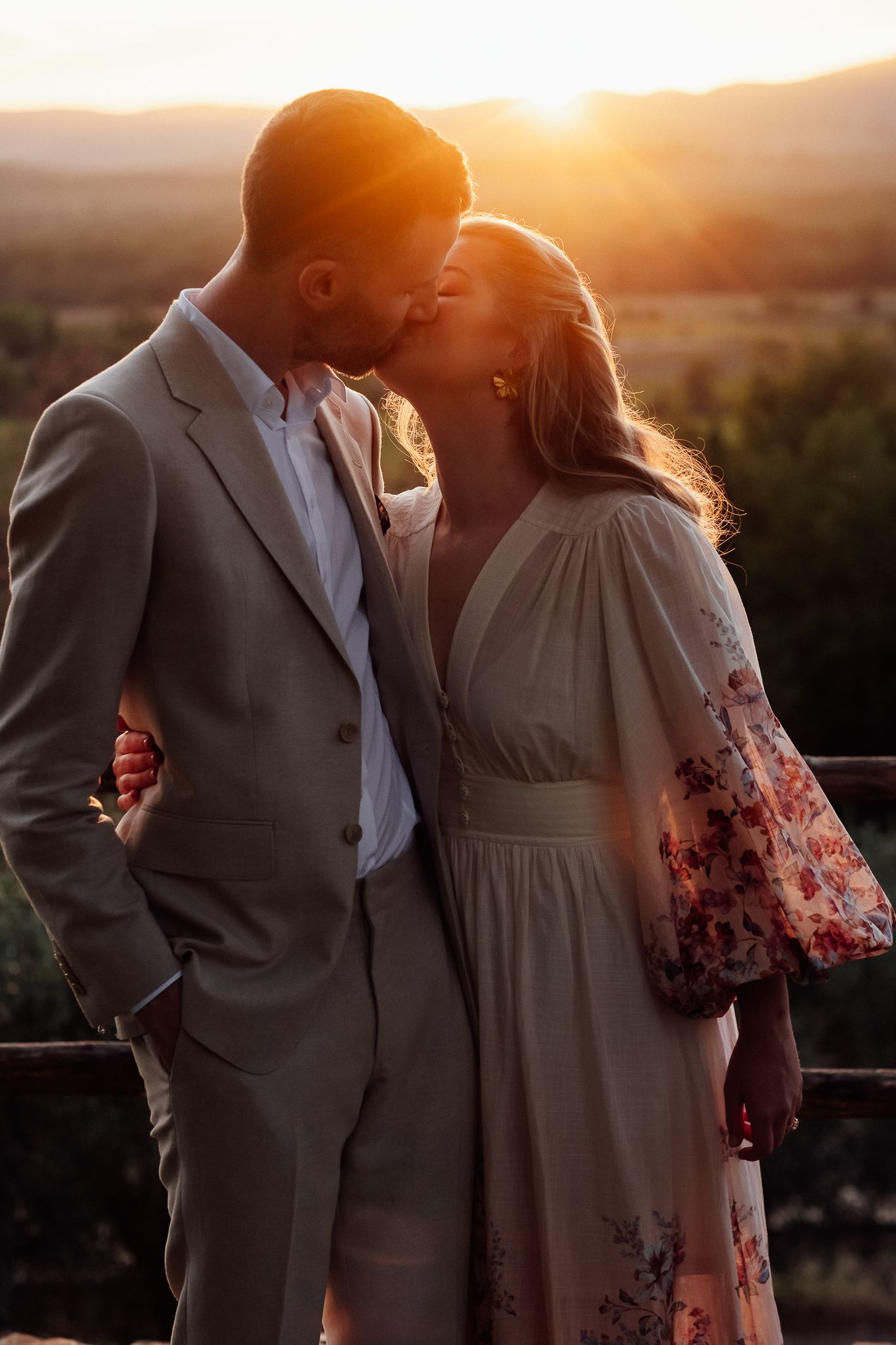 A couple kisses at golden hour in a countryside setting, he in a tan suit and she in a floral puff-sleeve dress.