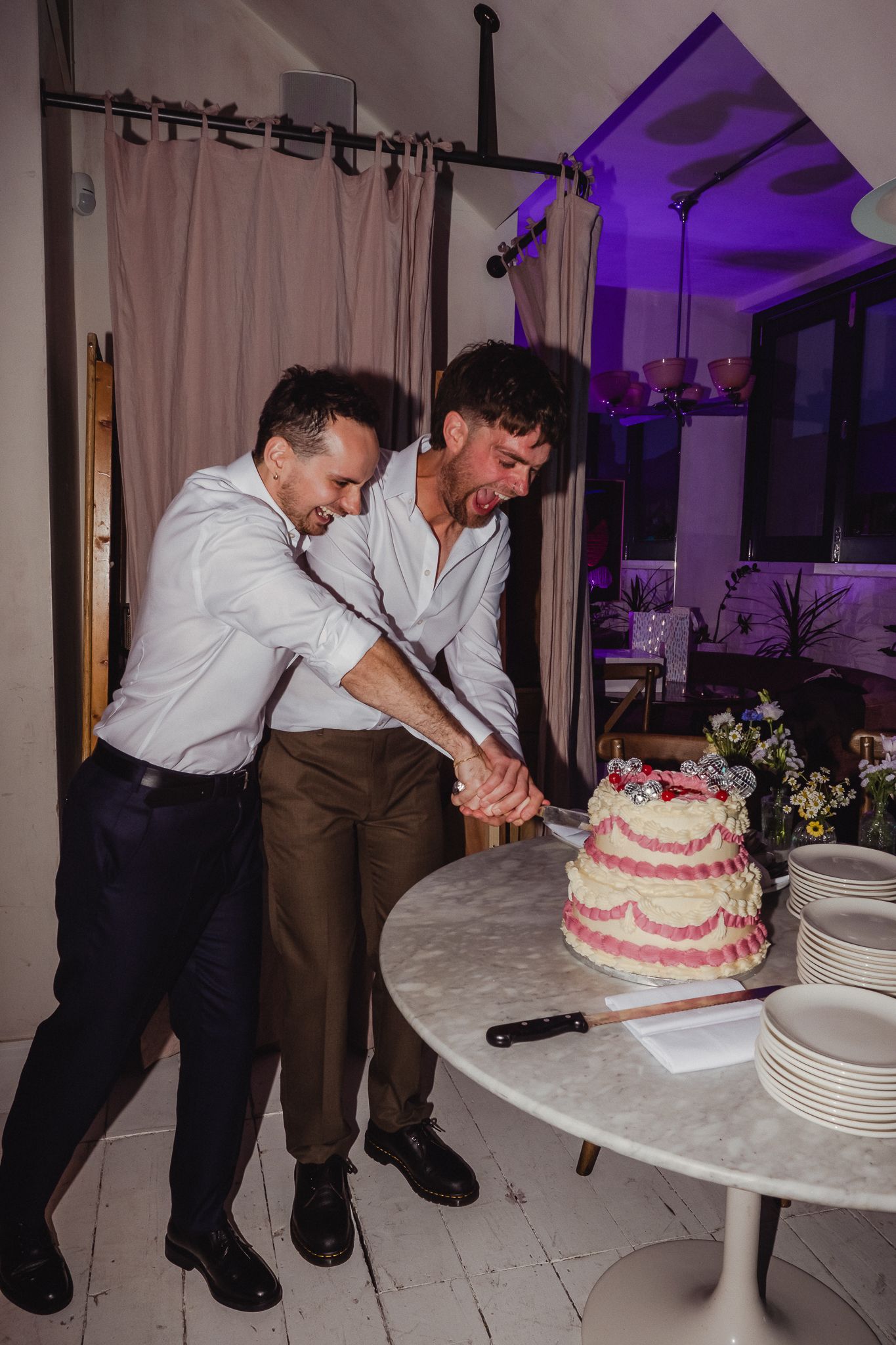 Two people cutting a tiered white and pink wedding cake together at a reception with purple lighting in the background.