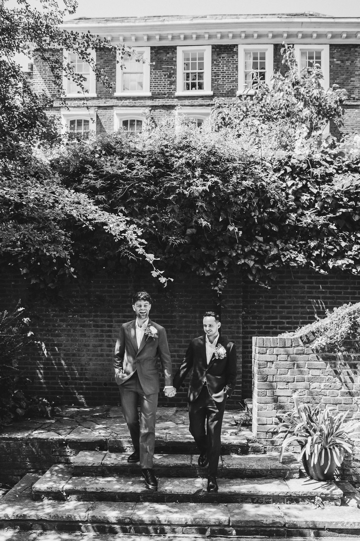A black and white photograph showing figures in formal attire standing against a brick wall with overhanging foliage.