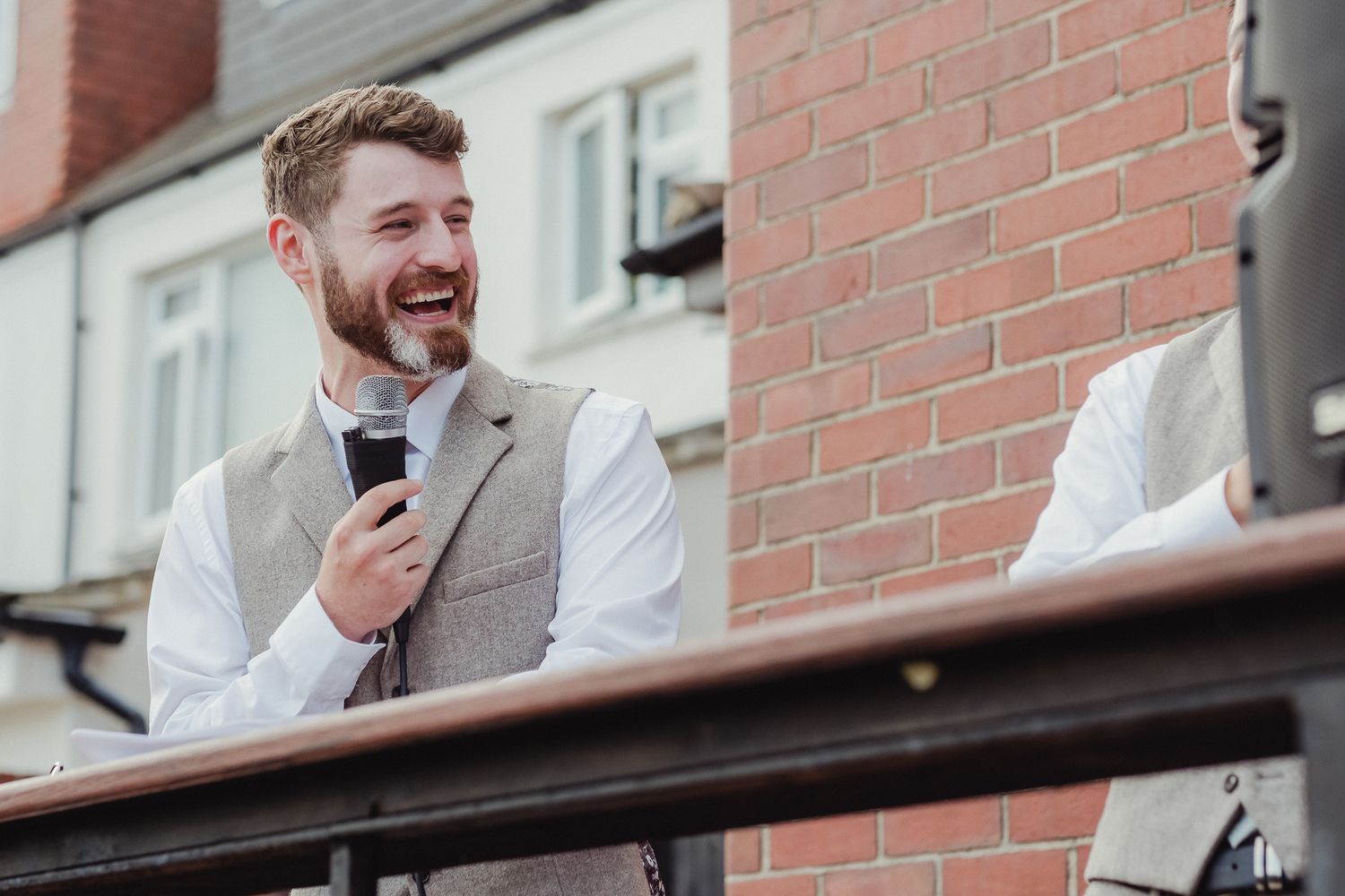 A group of people in formal attire laugh and smile while standing against a red brick wall.