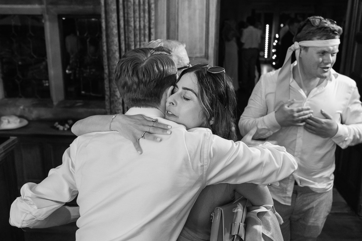 Black and white photo of two people sharing a warm hug at a social gathering, with guests visible in background.