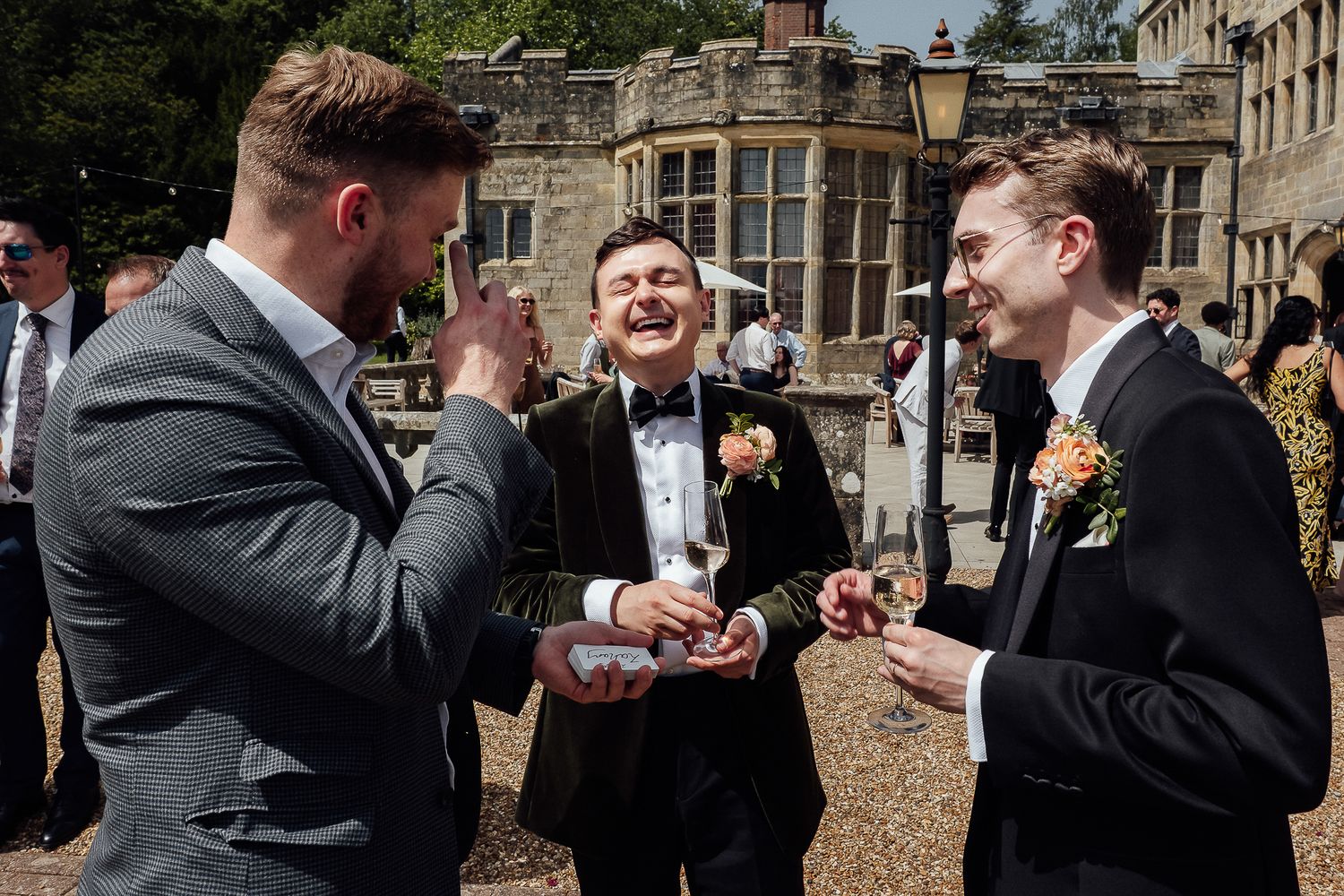 Three men in formal suits laughing and celebrating outdoors at a wedding reception on a sunny day.