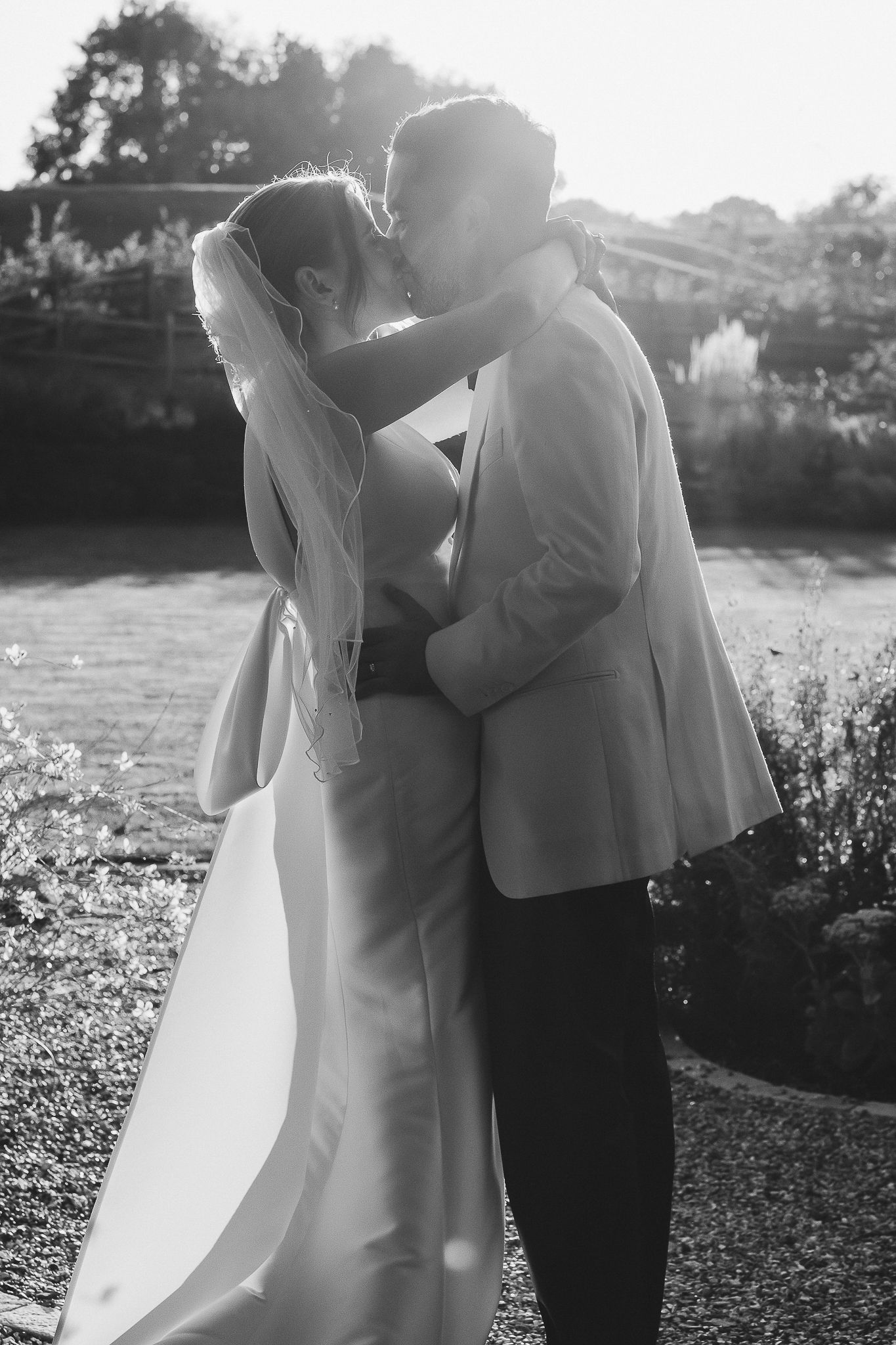A couple in wedding attire share an intimate embrace outdoors during sunset in a garden setting.