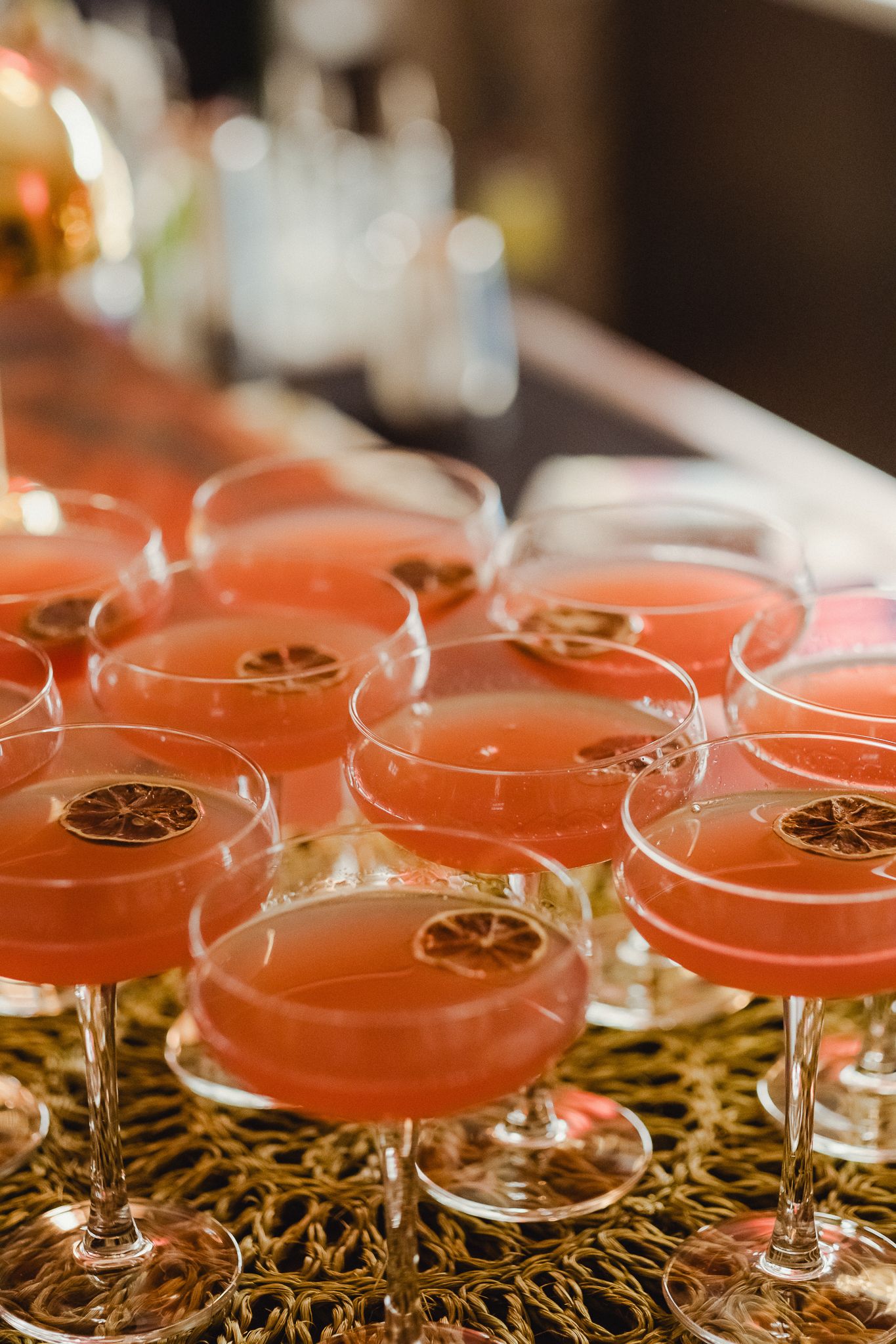 Multiple pink cocktails garnished with citrus slices are lined up on a decorative gold surface in a bar setting.