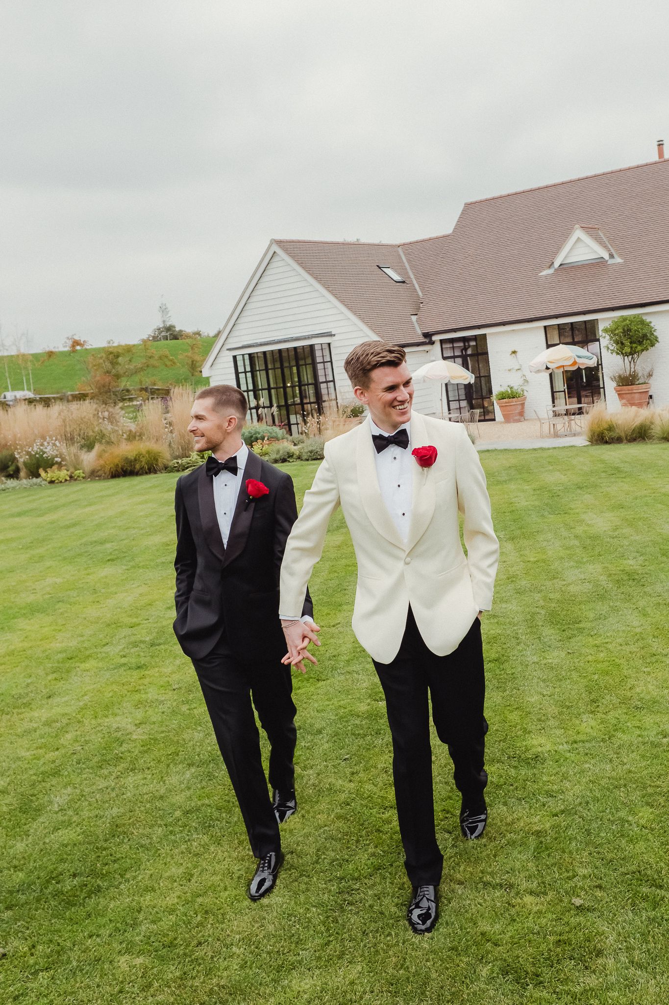 Two people in formal attire walk together and smile on a green lawn with houses visible in the background.